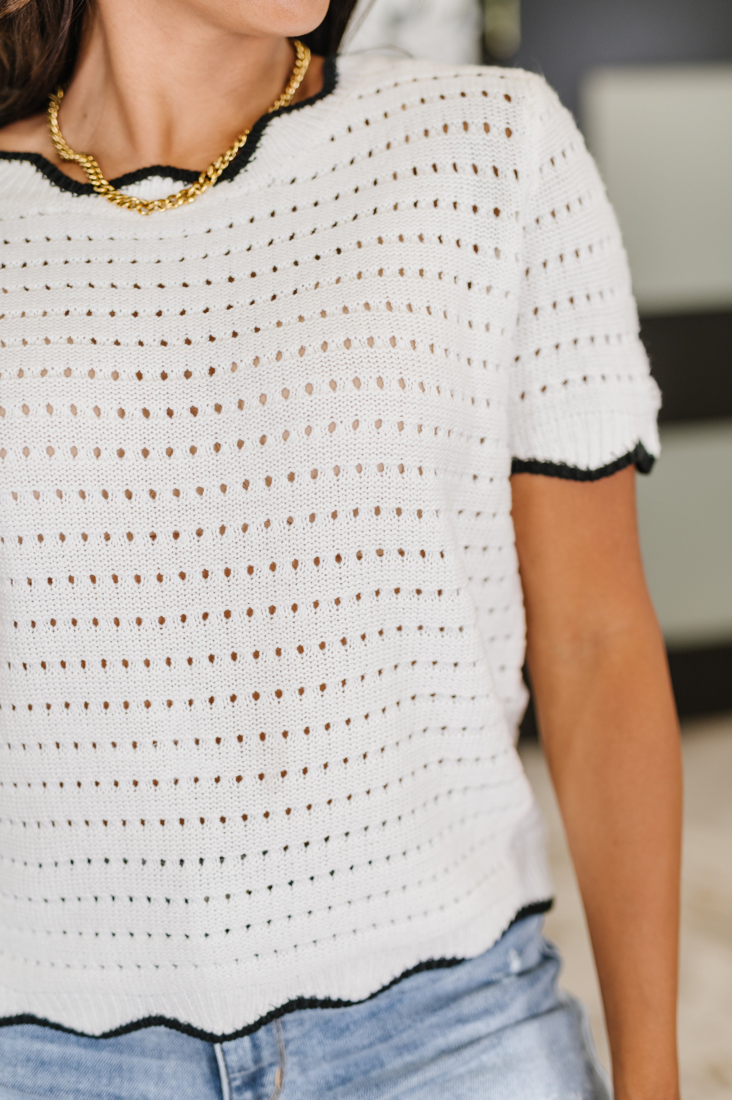 Close-up of wavy eyelet knit top in white with black trim, styled with gold chain necklace and casual denim.
