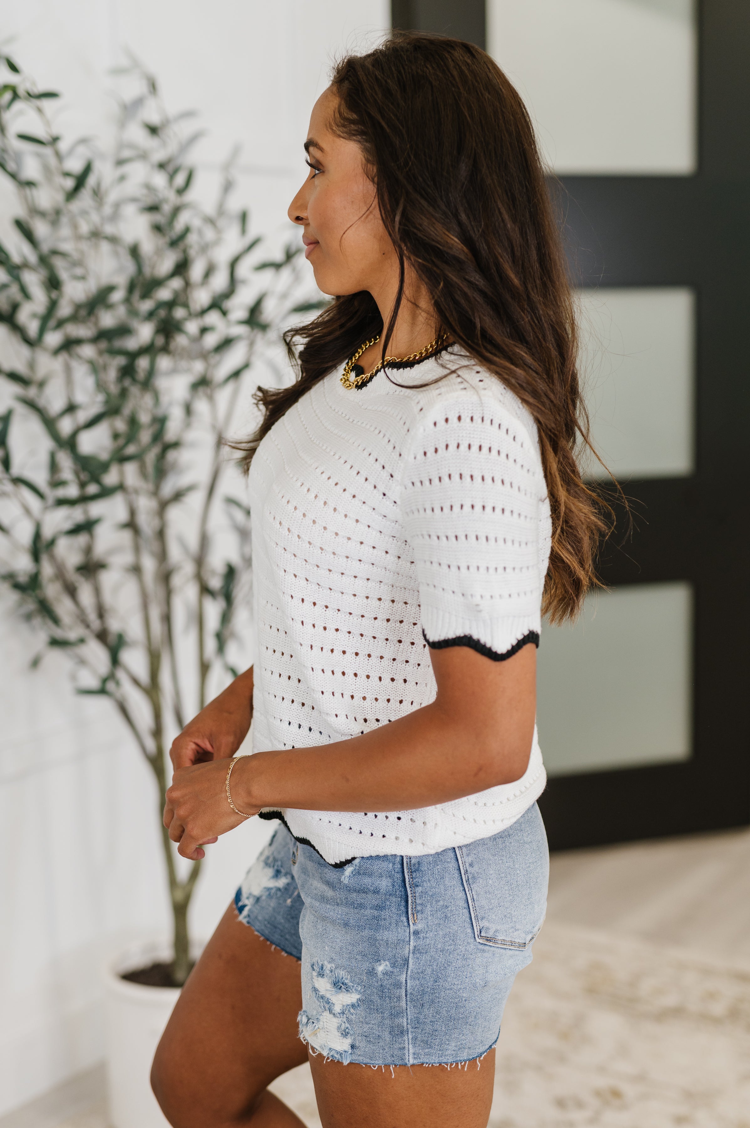 Woman wearing a white wavy eyelet knit top with black trim paired with distressed denim shorts indoors, eclectic style.