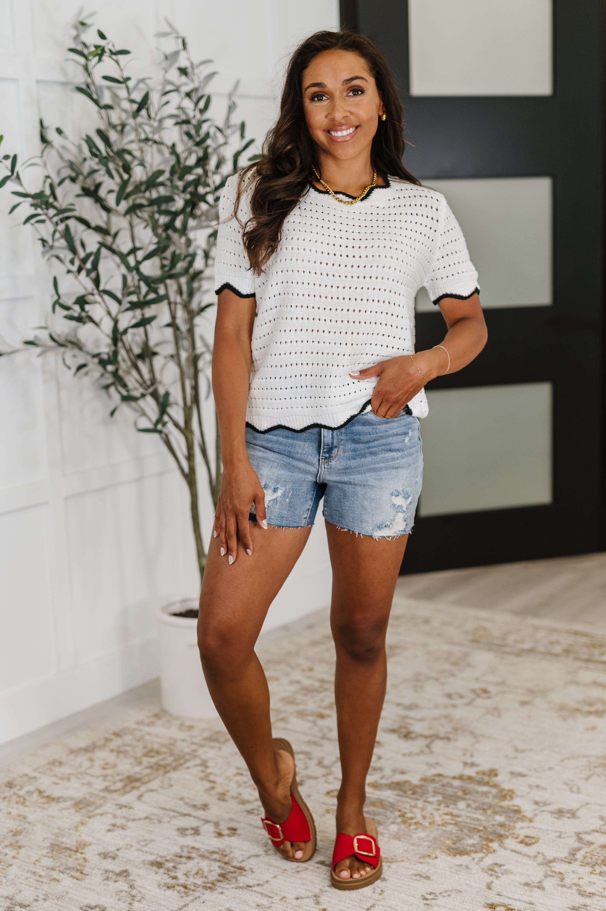 Woman wearing a white eyelet knit top with wavy black trim paired with distressed denim shorts and red sandals indoors