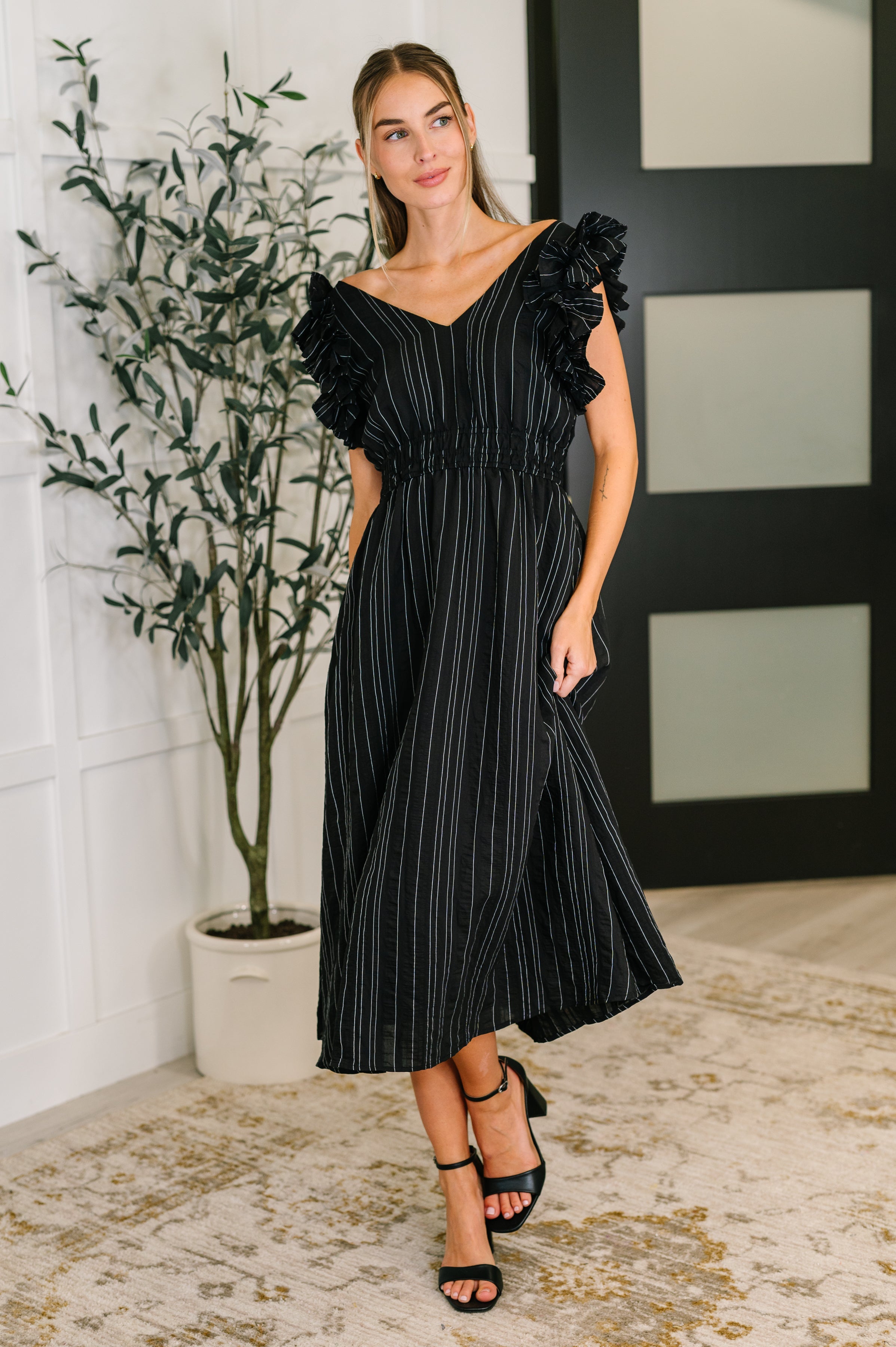 Woman wearing a black pinstripe dress with ruffled cap sleeves and V-neck posing indoors near a plant