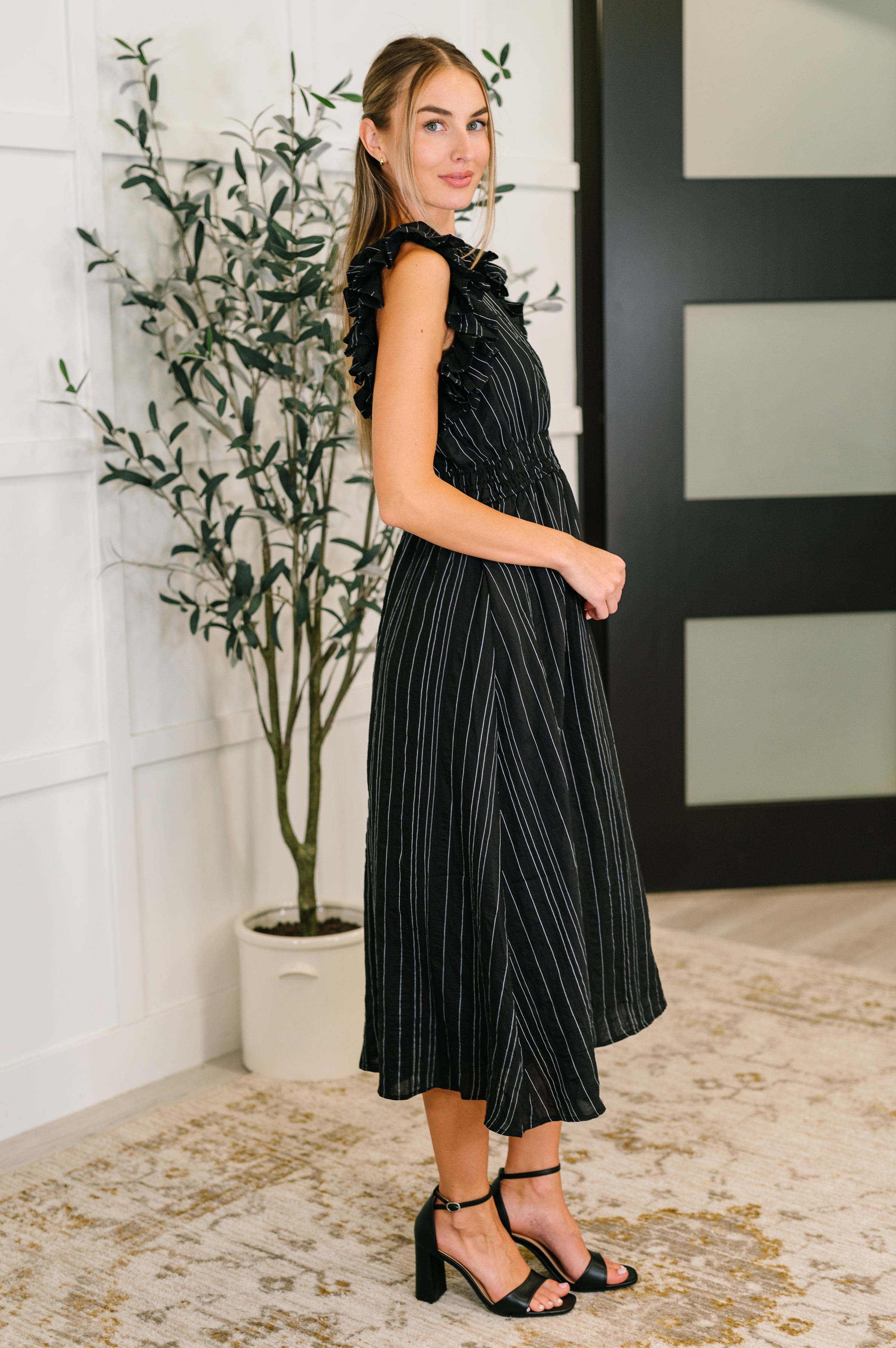 Profile of woman in black pinstripe dress with ruffled sleeves and midi length standing indoors by plant