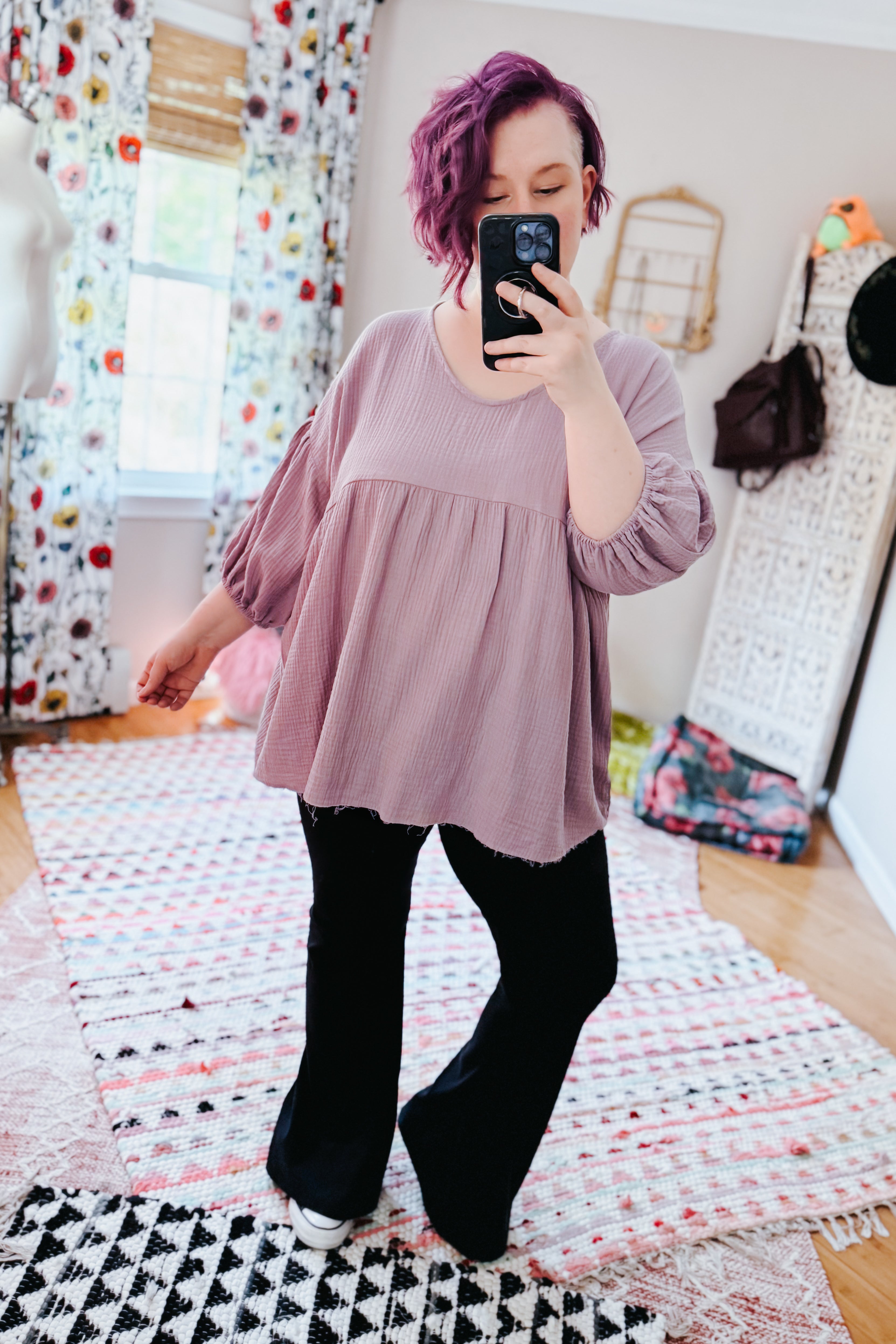 Woman with purple hair wearing a purple bubble sleeve peasant blouse and black pants in a bright, eclectic room