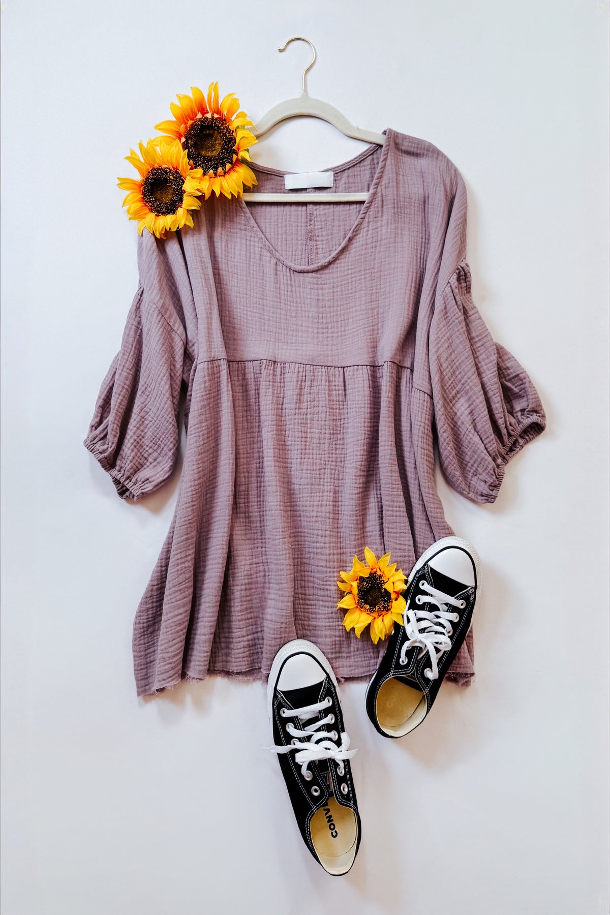 Purple cotton gauze peasant blouse with balloon sleeves and babydoll fit displayed on a hanger with sunflowers and black sneakers