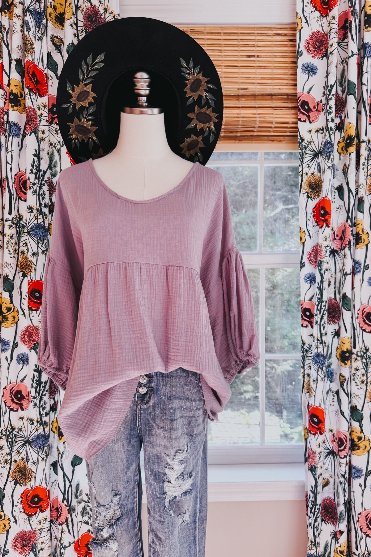 Purple cotton gauze peasant blouse with balloon sleeves and raw hem on a mannequin styled with distressed jeans and a wide brim hat