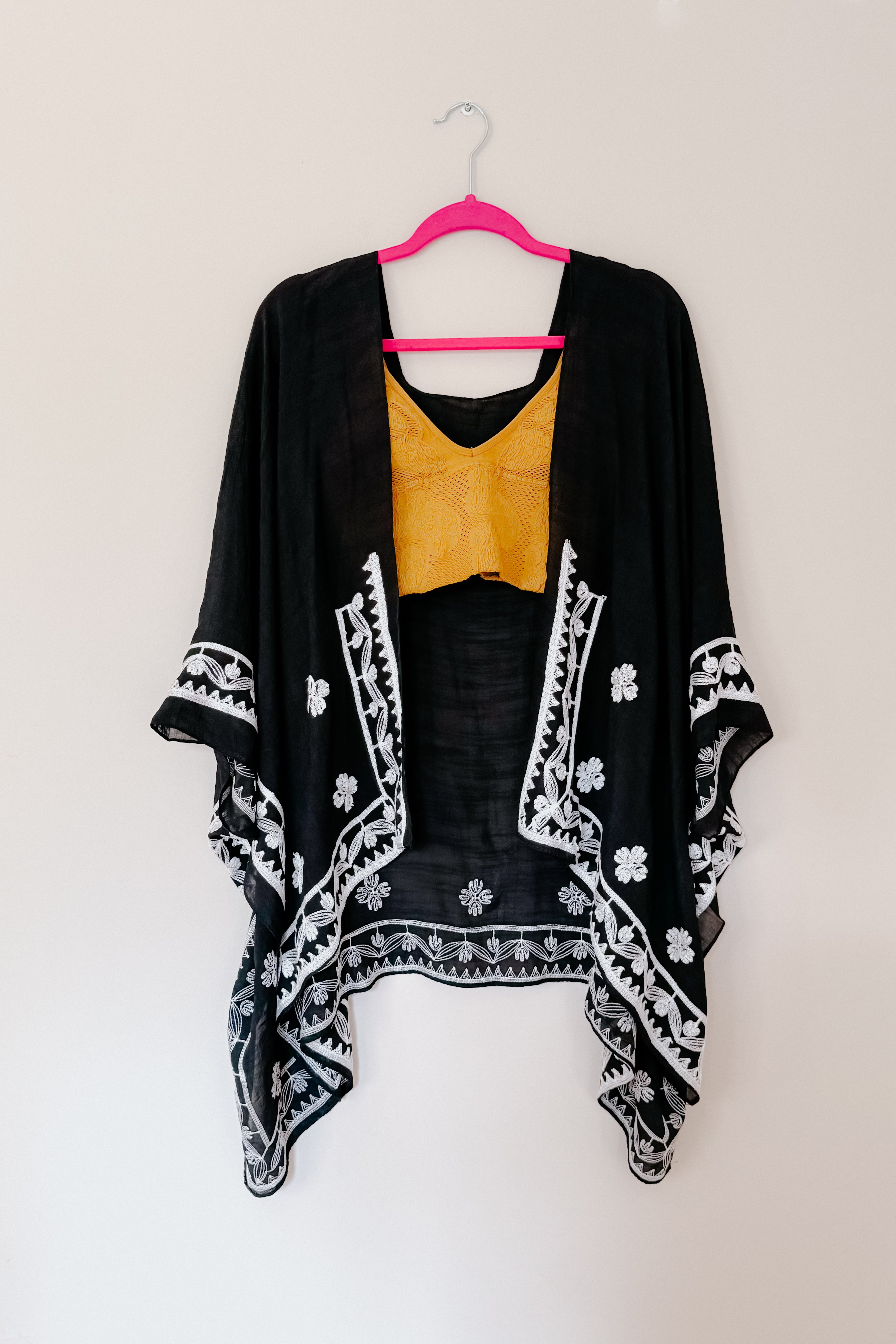 Black embroidered layering piece with white floral details hanging over mustard textured top on pink hanger in eclectic style