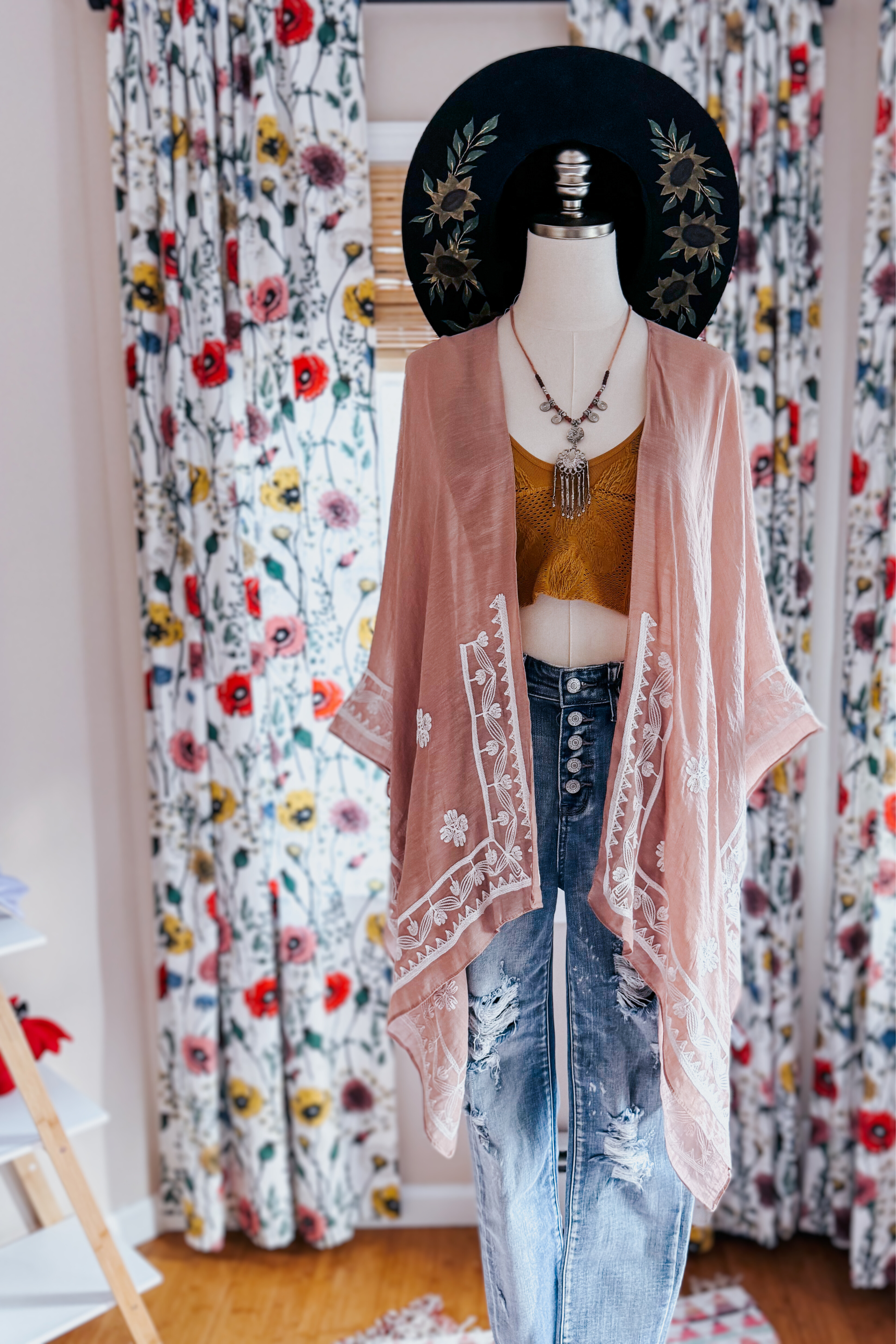 Mannequin dressed in blush taupe embroidered layering piece over mustard top and distressed jeans with black hat indoors