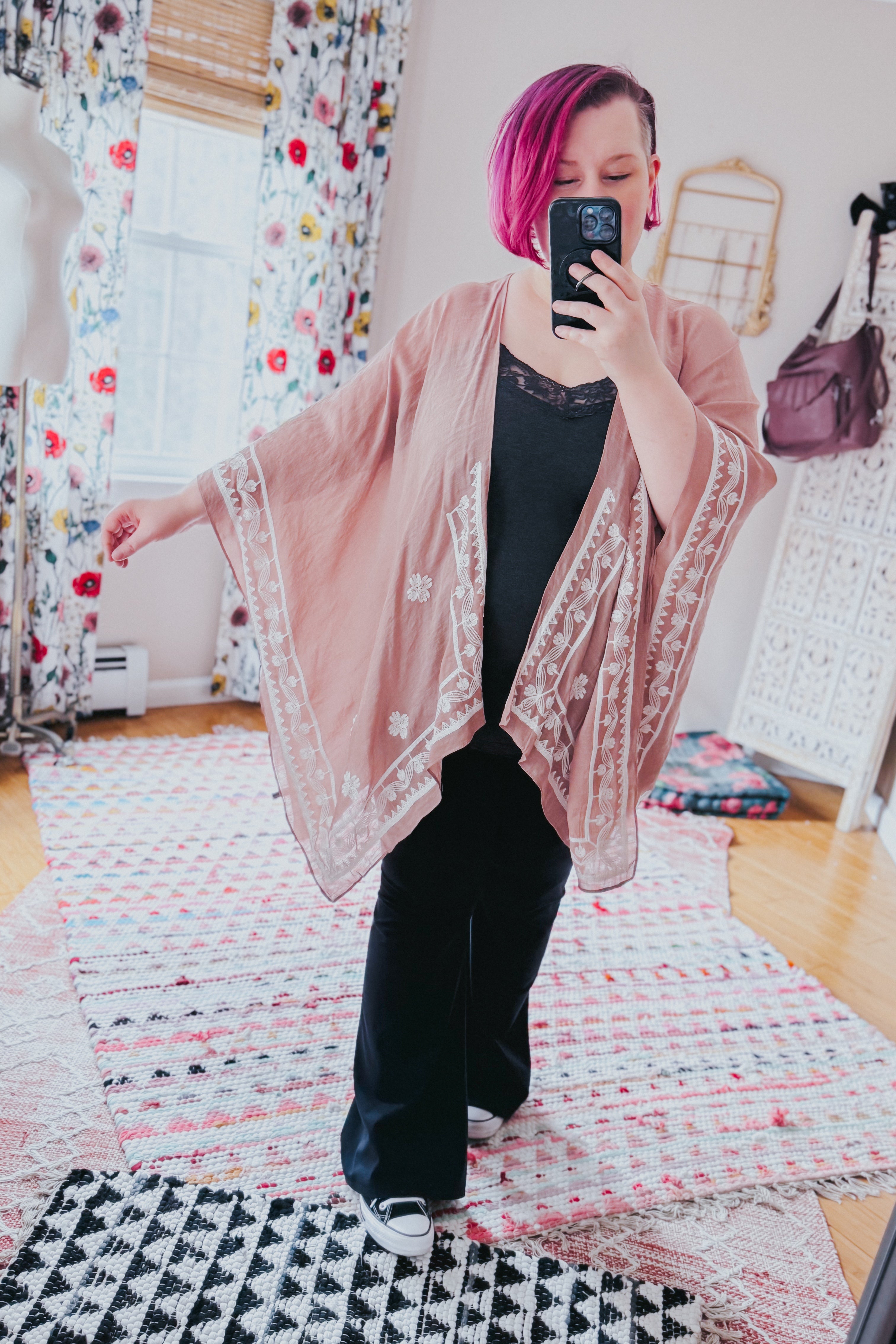 Woman with vibrant magenta hair wearing a blush taupe layering piece with white embroidery over black outfit indoors