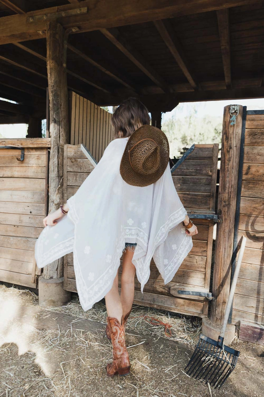 Woman wearing sheer stone layering piece with embroidered trim and cowboy boots holding wide-brimmed hat outdoors