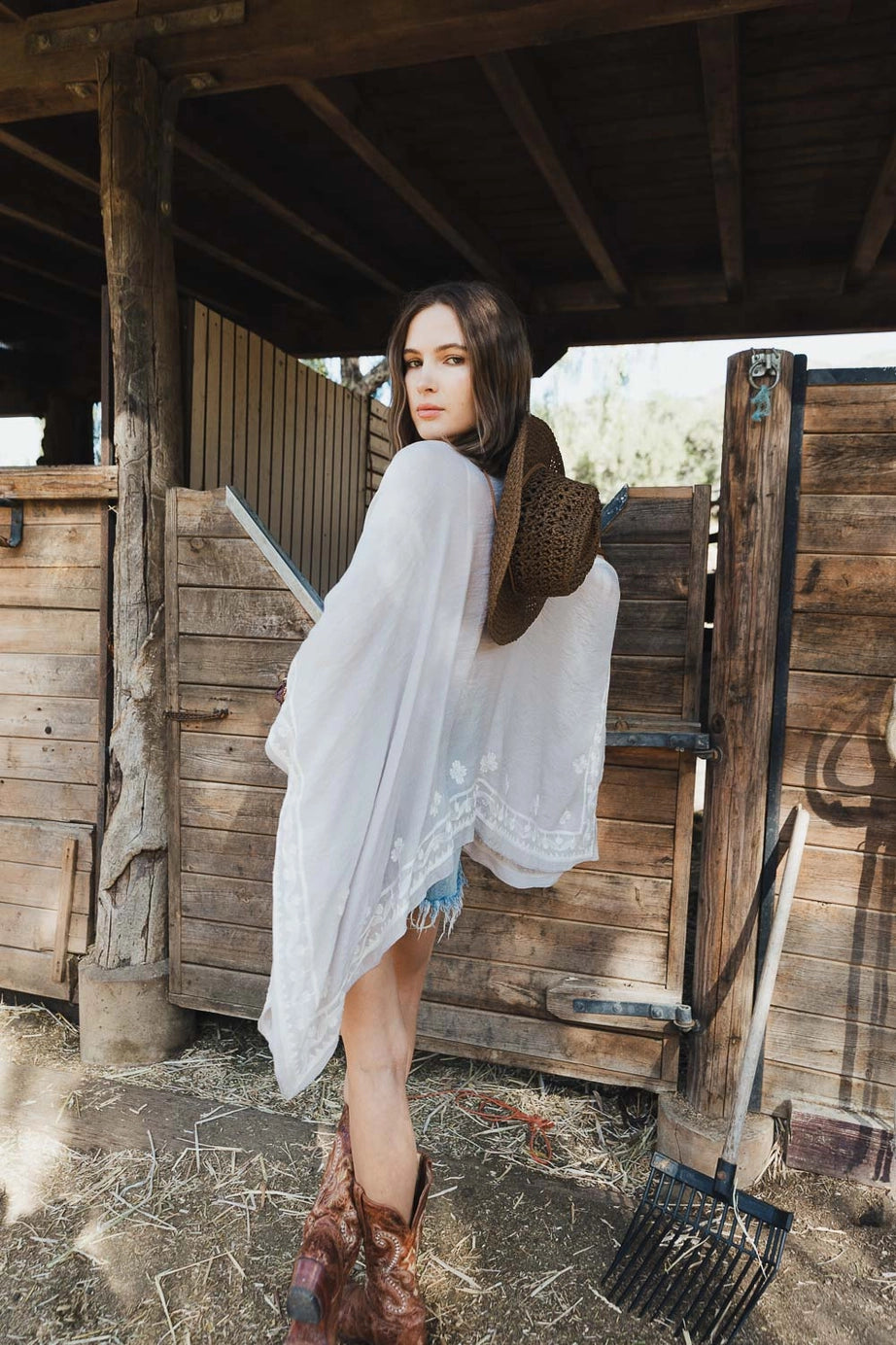 Woman standing in rustic barn setting wearing flowing stone layering piece with subtle embroidery and cowboy boots holding hat