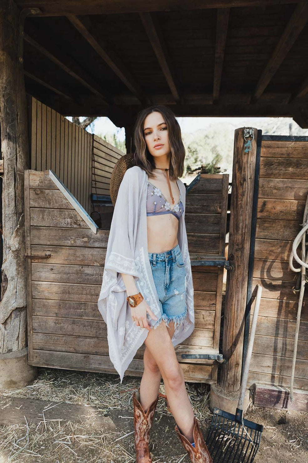 Woman in stone-colored embroidered layering piece over floral bralette and distressed denim shorts standing in rustic wooden barn setting