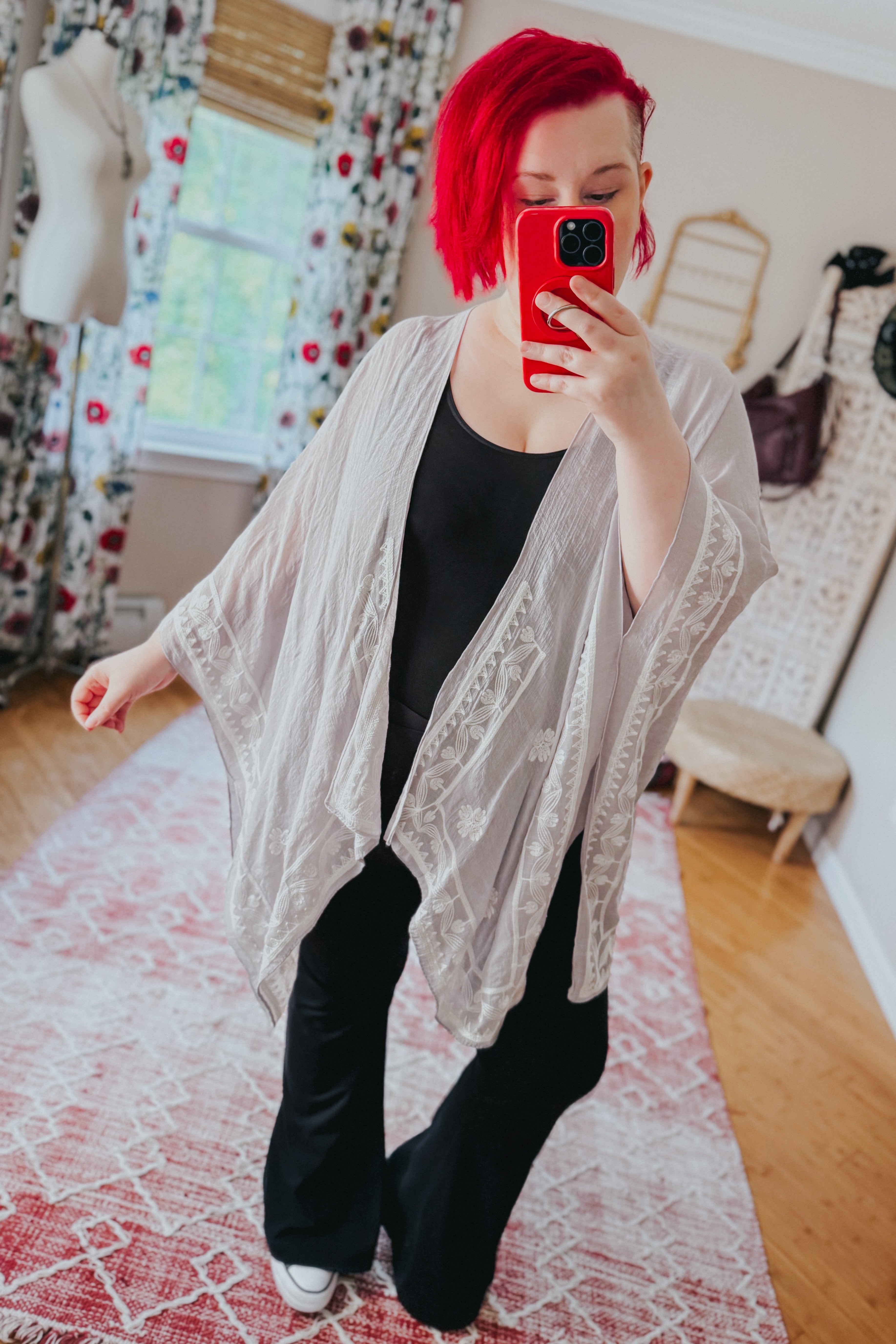 Woman with bright red hair wearing flowy stone layering piece with subtle embroidery over black outfit indoors taking mirror selfie