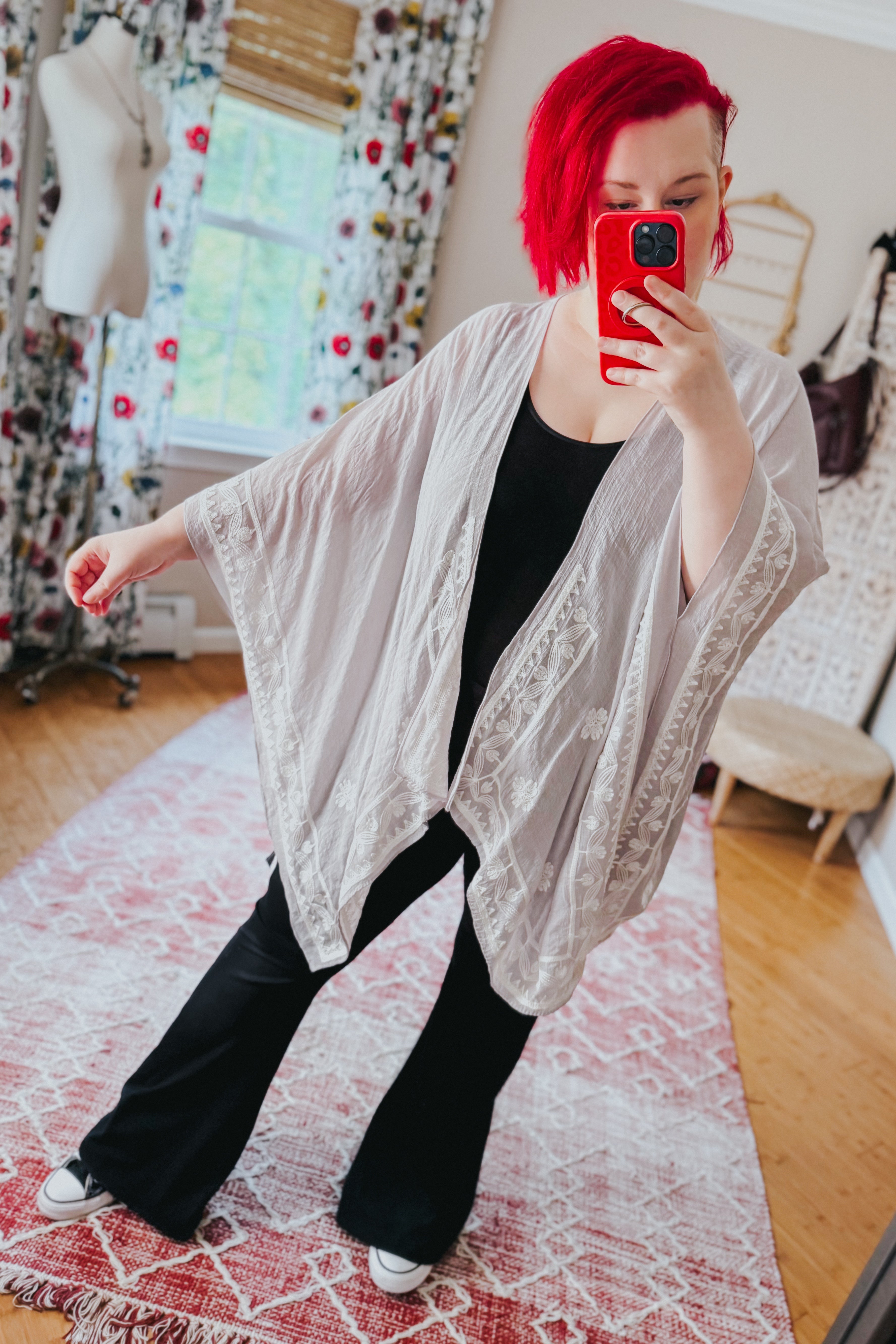 Woman with bright red hair wearing stone-colored embroidered layering piece over black outfit taking mirror selfie indoors
