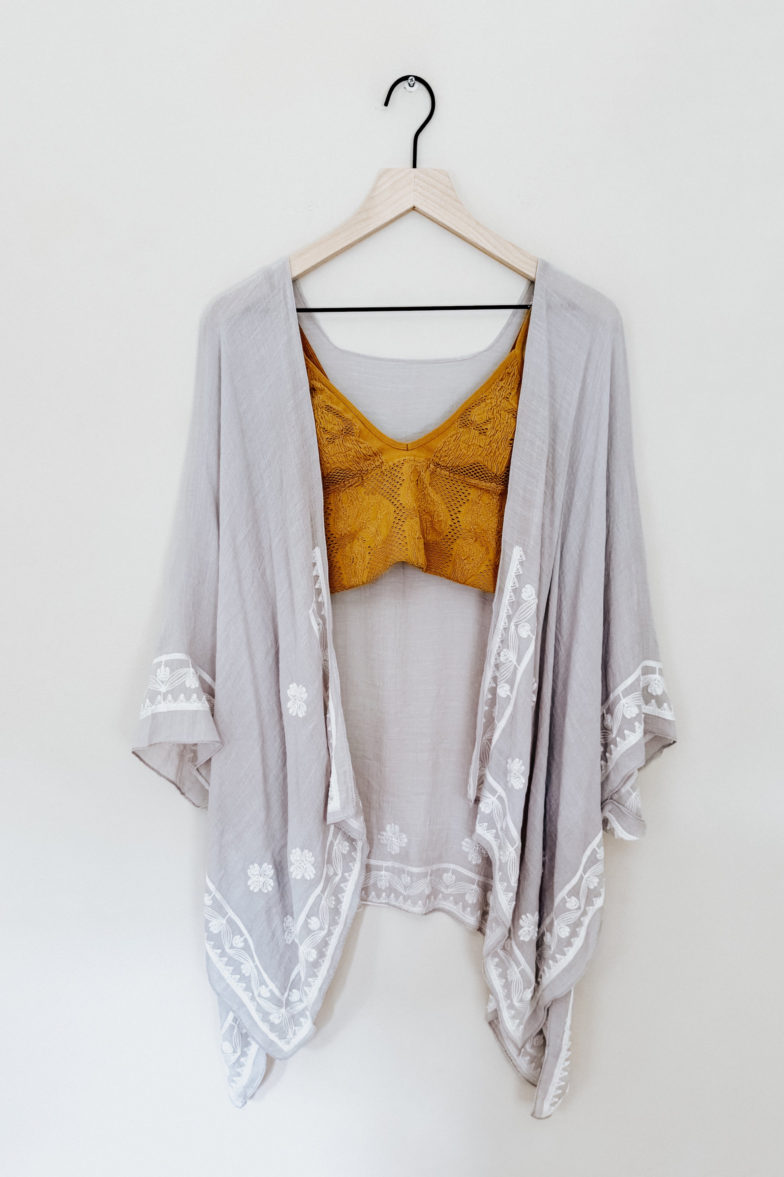 Stone-colored layering piece with white poppy embroidery draped over a mustard textured crop top on wooden hanger