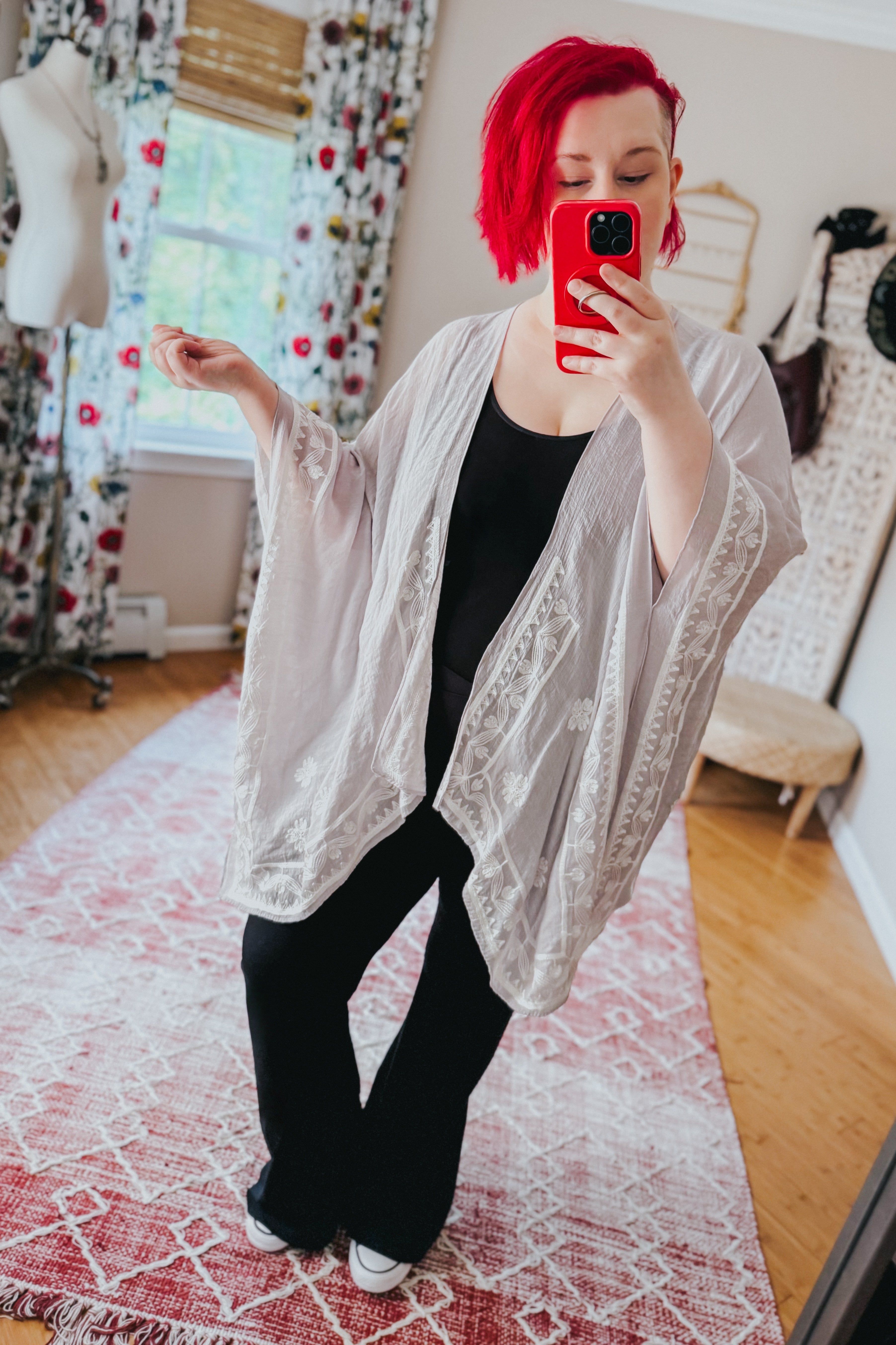 Woman with bright red hair wearing stone-colored embroidered layering piece over black outfit indoors taking mirror selfie