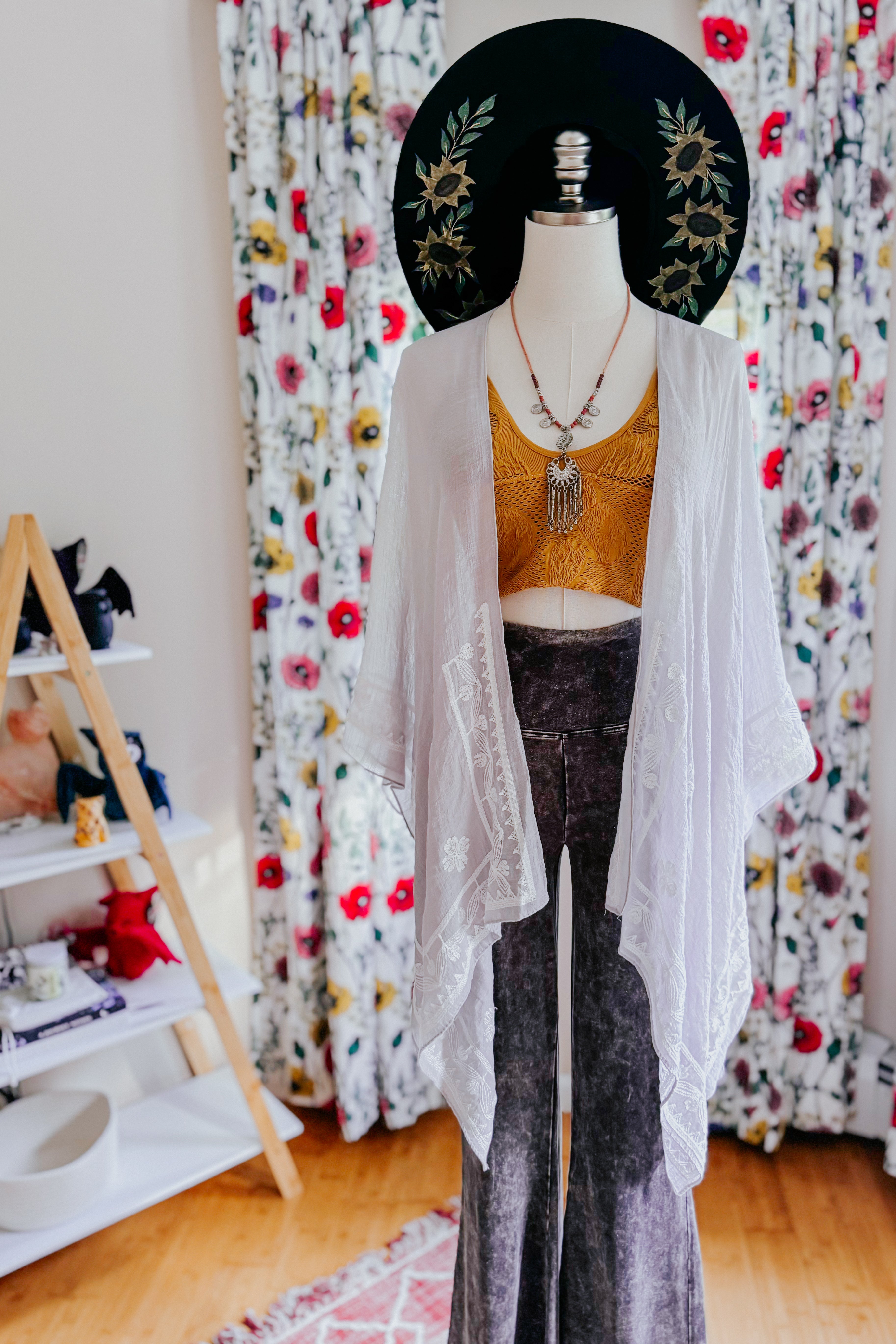 Mannequin dressed in sheer stone layering piece with delicate embroidery, mustard crop top, gray velvet pants, and embroidered black hat indoors