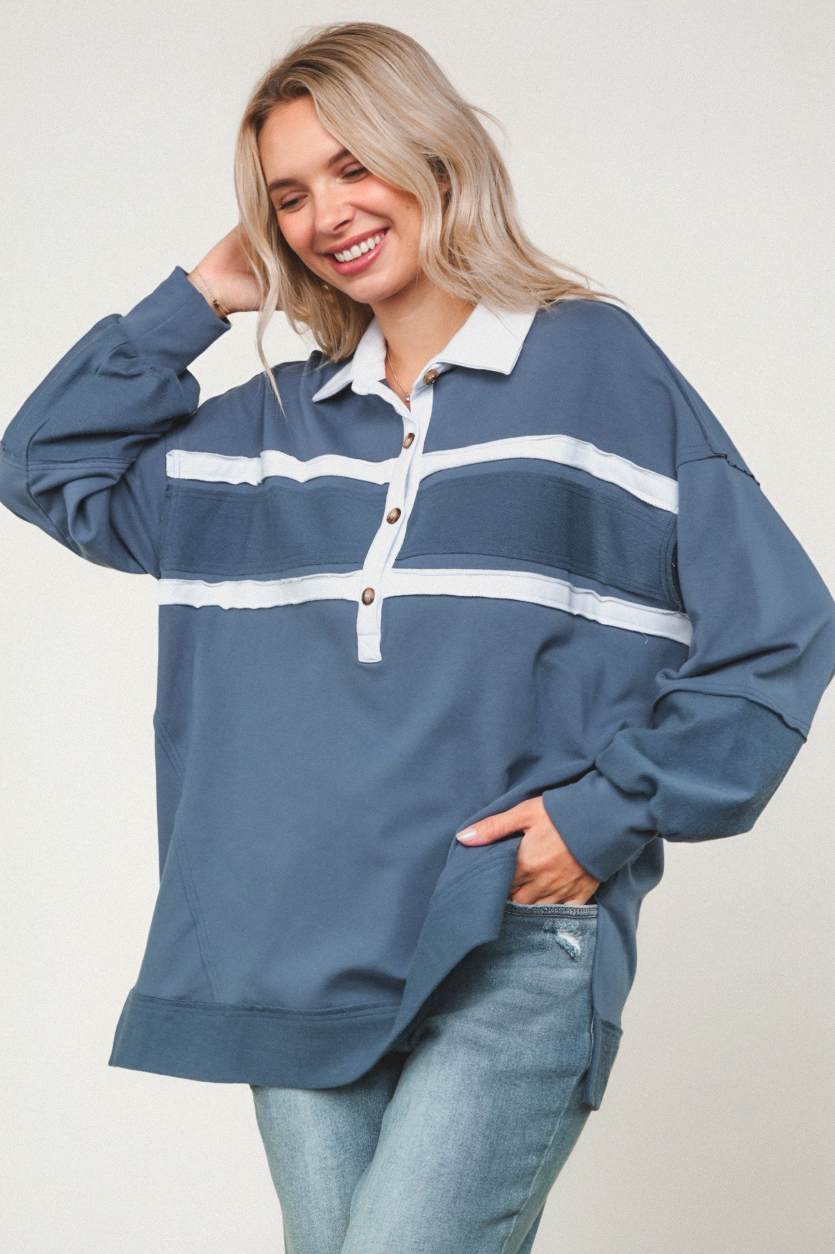 Smiling woman wearing oversized steel blue polo top with white stripes and wooden buttons paired with light washed jeans