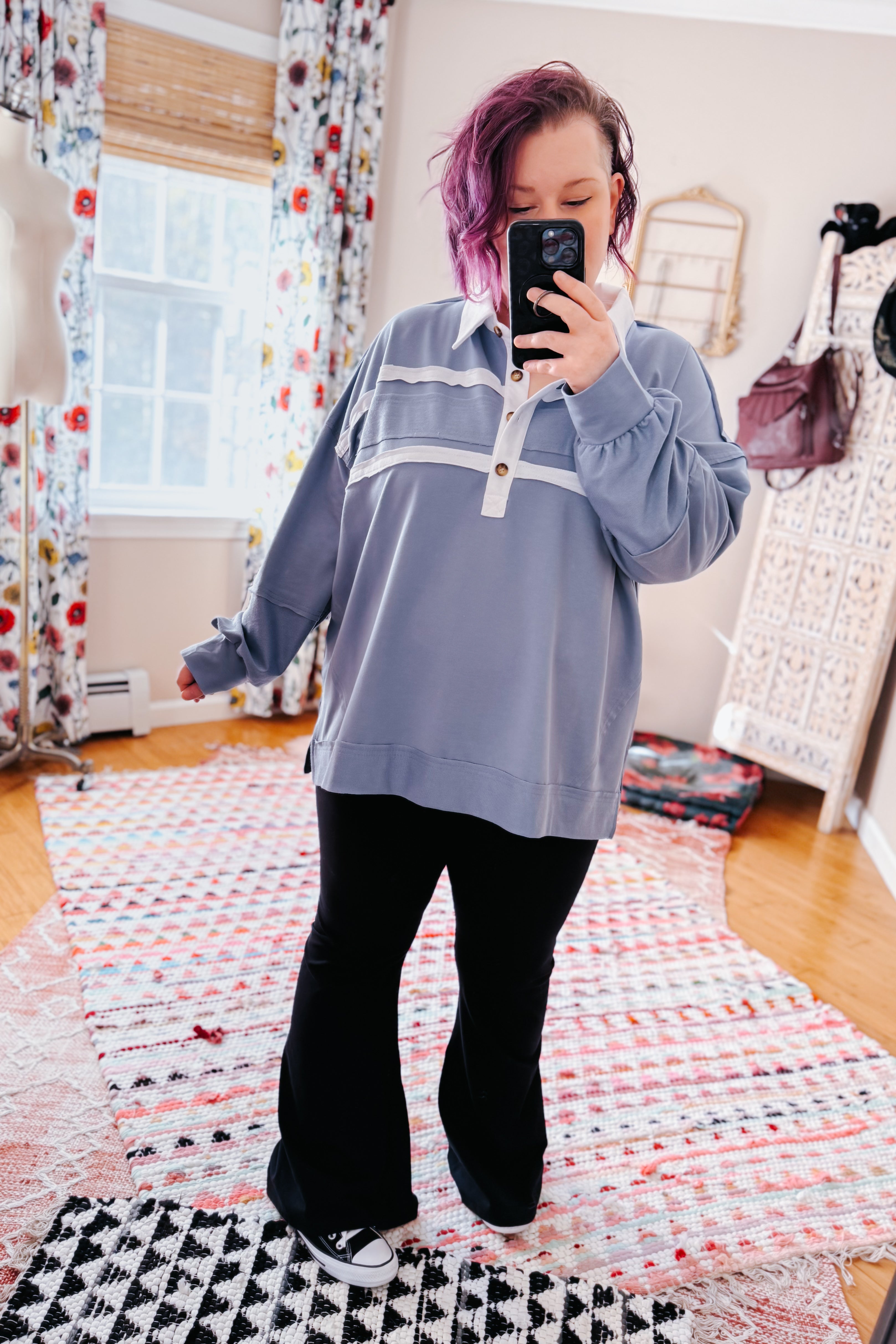 Woman wearing relaxed slate blue polo top with white collar and stripes paired with black flared pants and black sneakers taking selfie