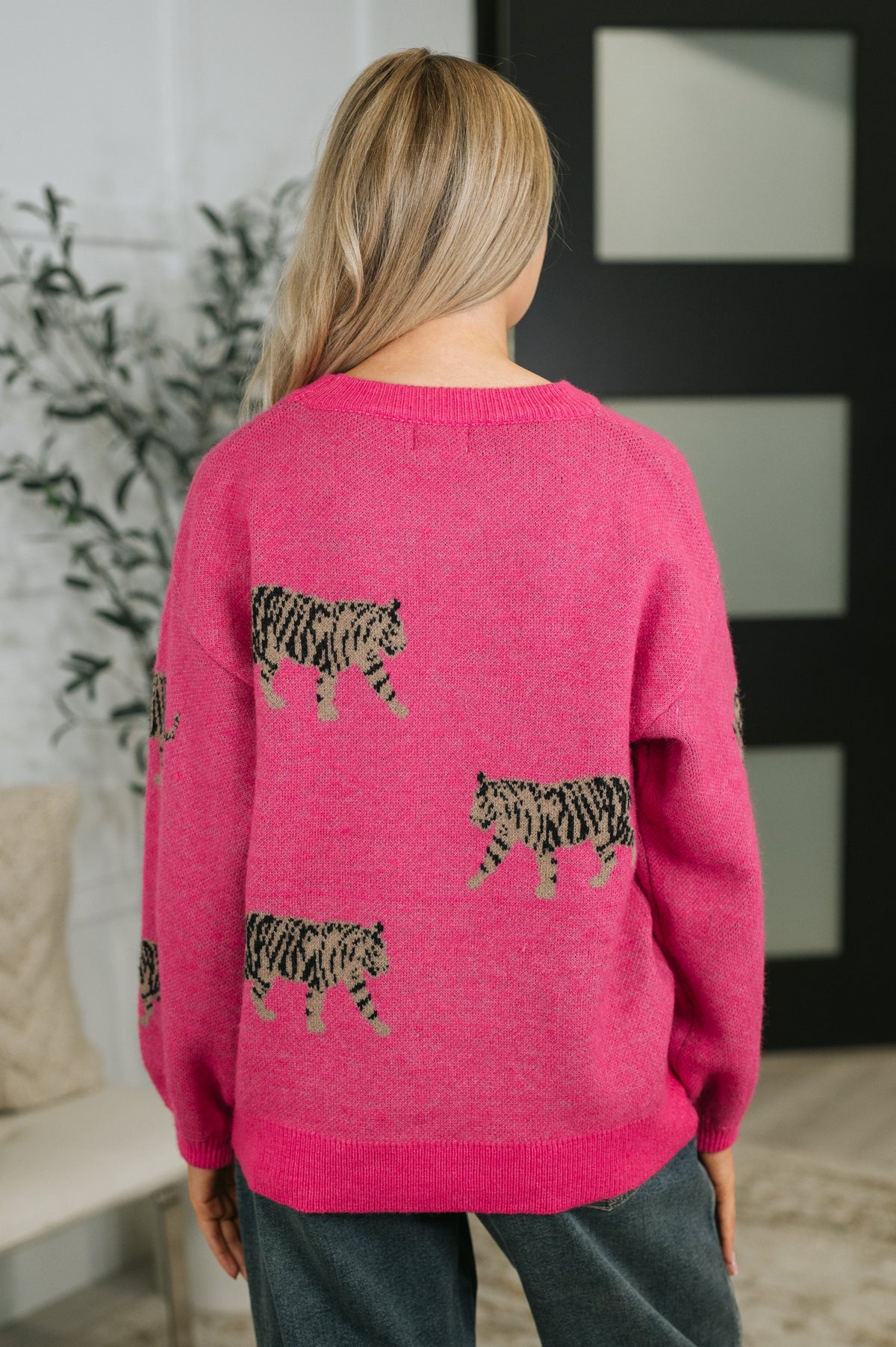 Back view of a woman wearing a pink knit pullover with scattered prowling tiger graphics and ribbed trim styled with dark jeans indoors