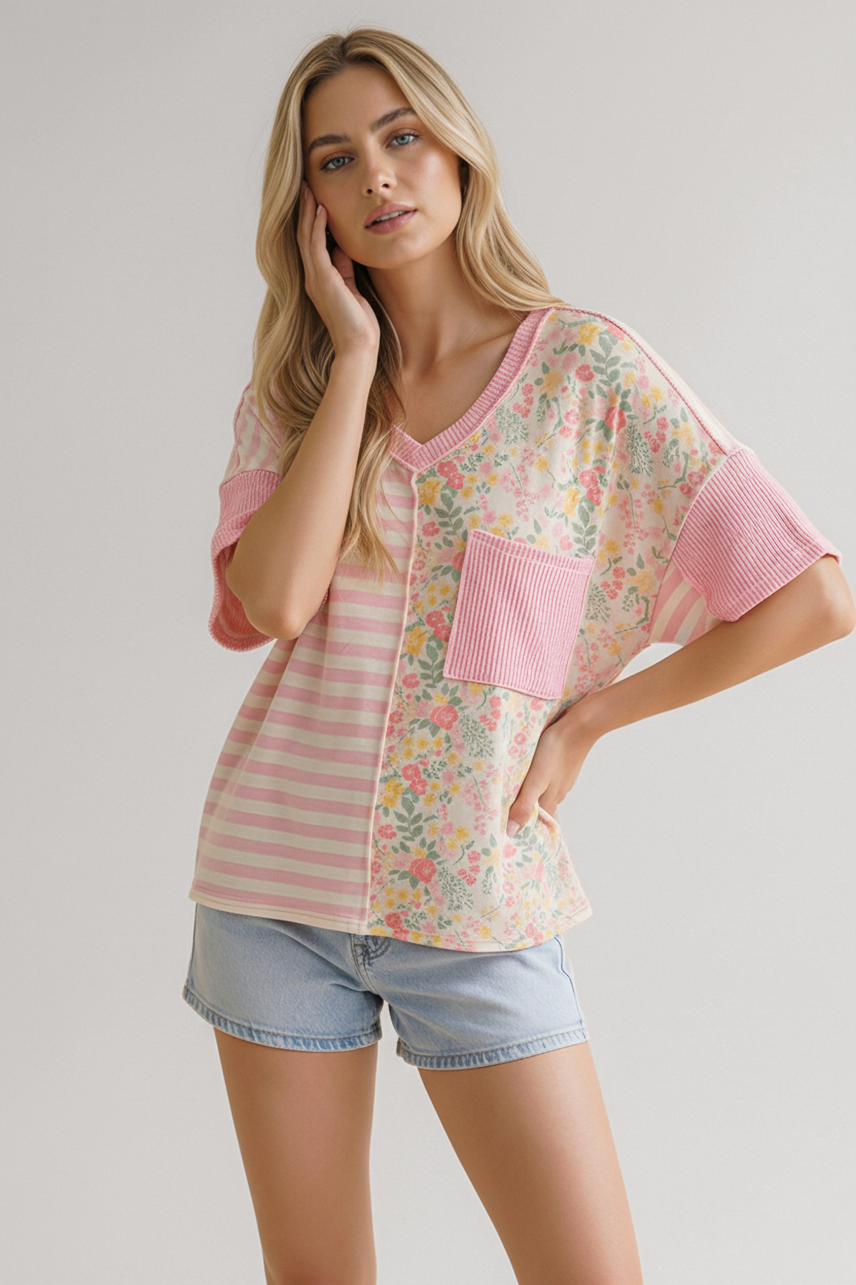 Woman wearing a pink top with floral and striped panels, a ribbed pocket, and rolled sleeves paired with light denim shorts