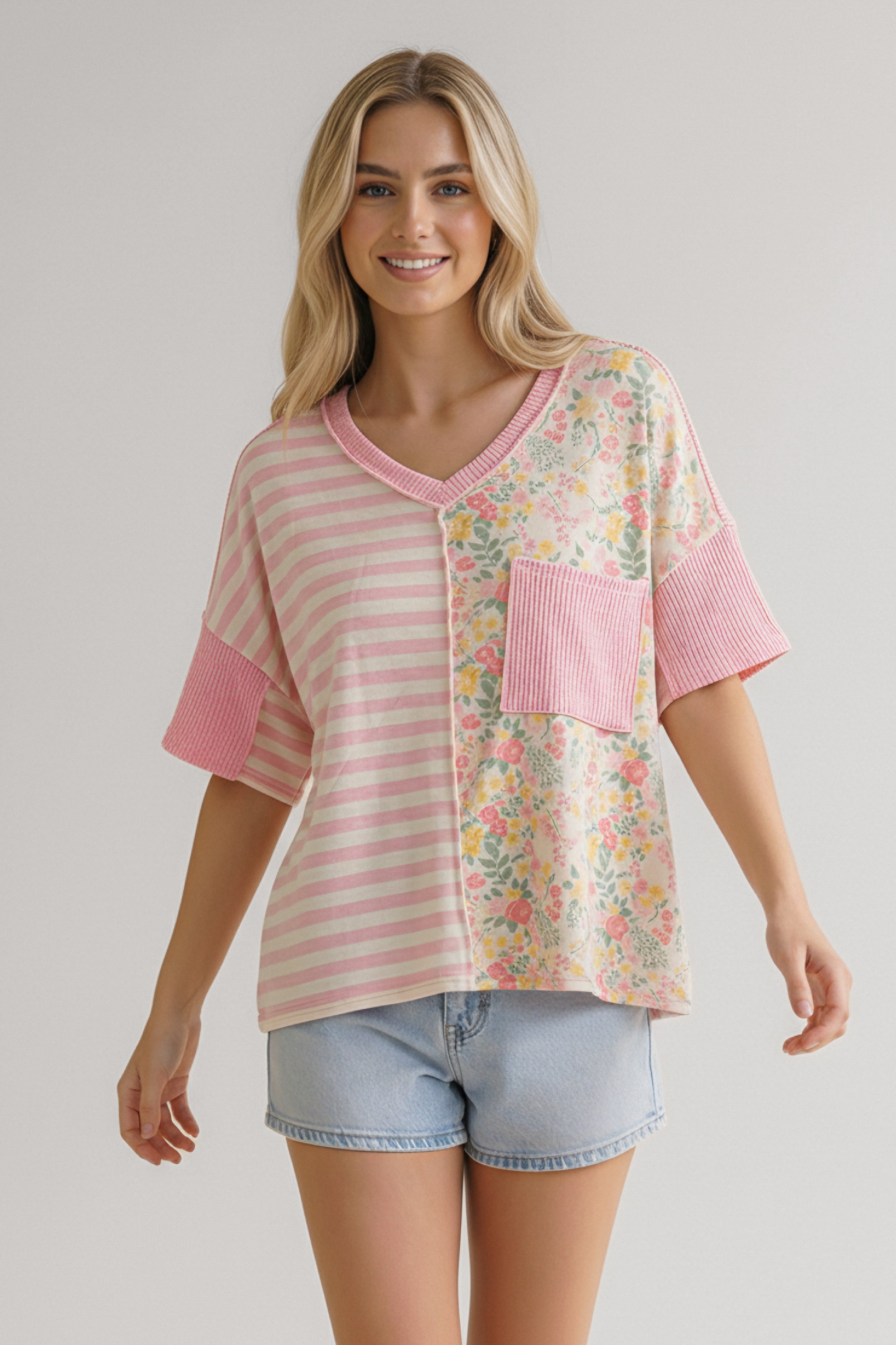 Woman smiling wearing a pink top with striped and floral panels, a ribbed pocket, and rolled sleeves paired with denim shorts