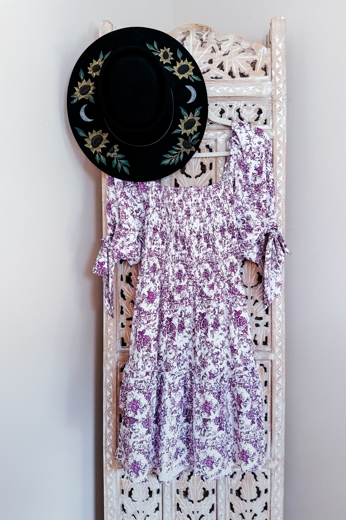 Purple floral babydoll dress with smocked bodice ruffled tiered skirt and sleeve ties displayed on an ornate white rack with a black hat