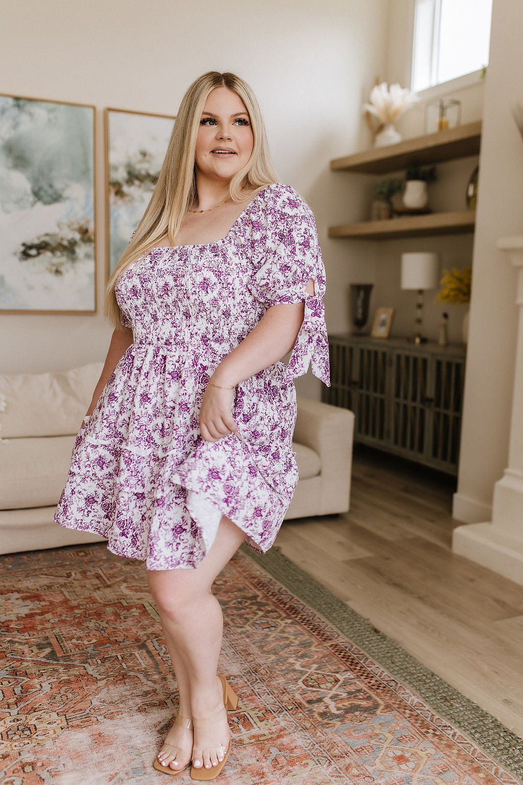 Plus size woman in purple floral dress with puff sleeves and square neckline in bright, eclectic home setting