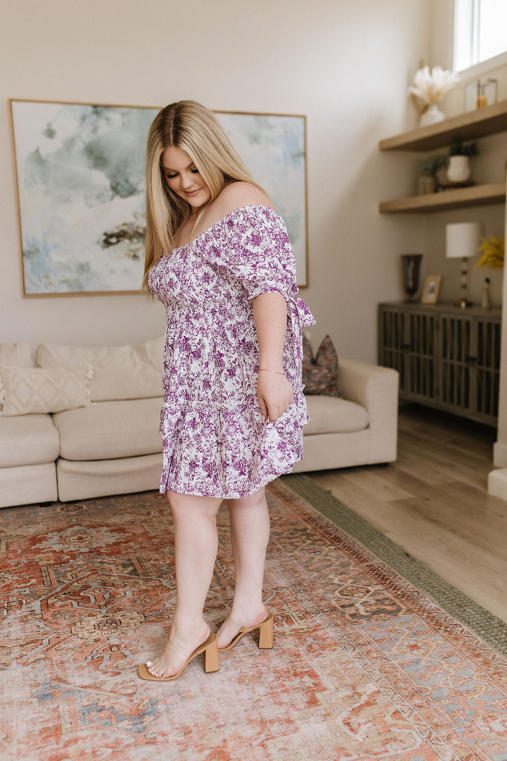 Plus size woman in purple floral off-shoulder dress with puff sleeves wearing nude block heel sandals in cozy living room