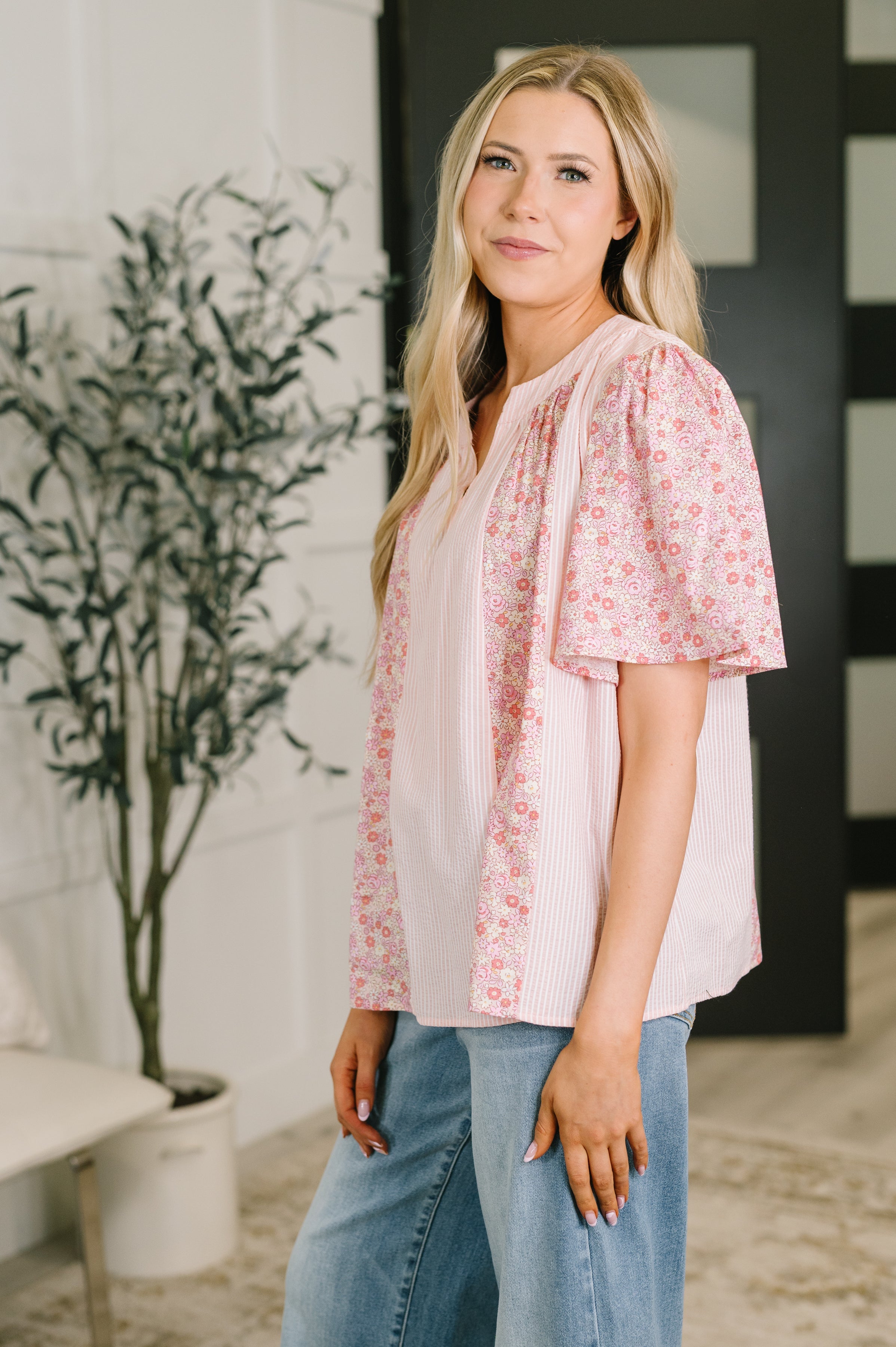 Side view of woman wearing a pink floral bell sleeve blouse with vertical stripes paired with blue jeans indoors