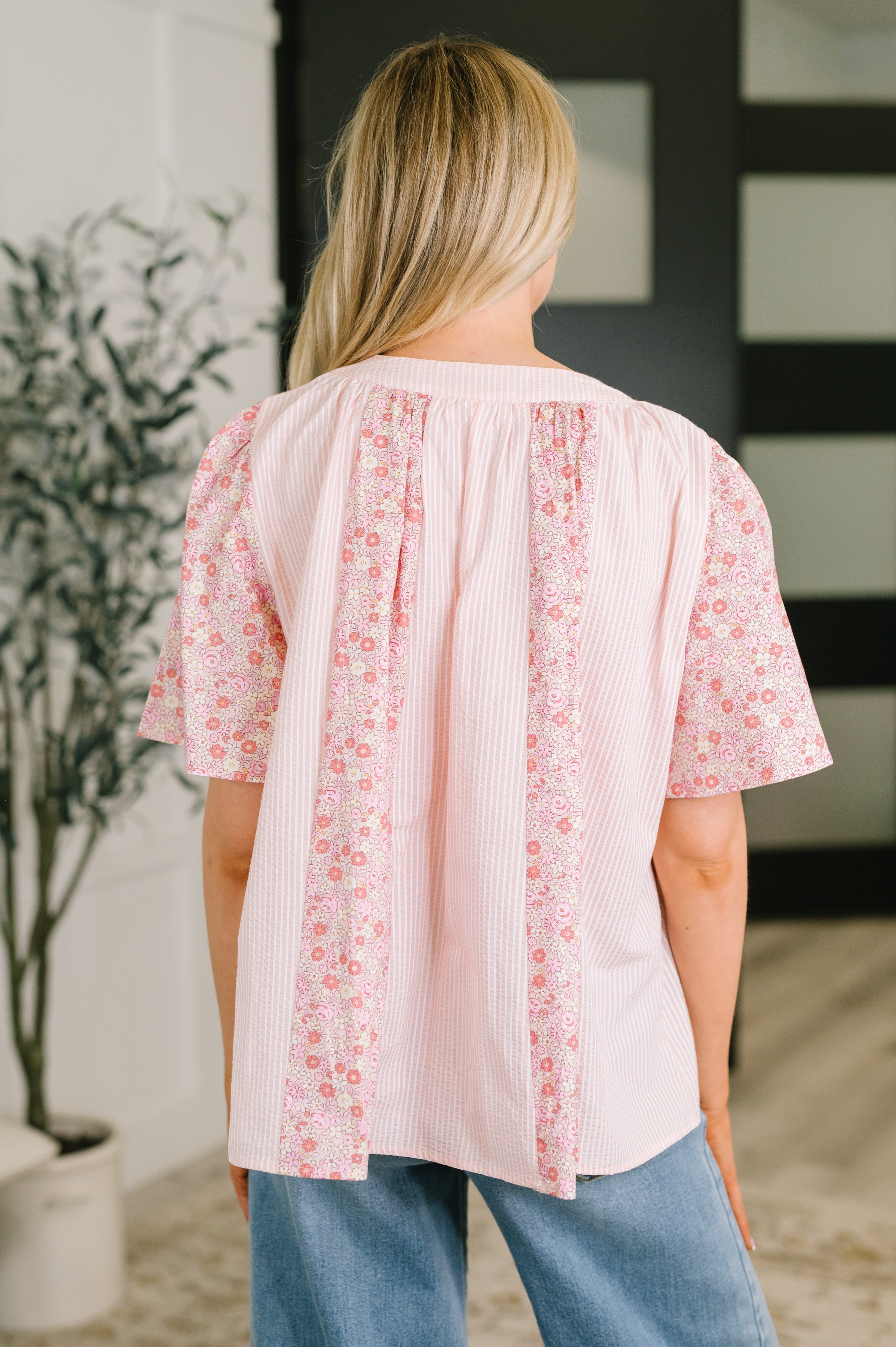 Back view of woman wearing a pink color block blouse with floral bell sleeves paired with light blue jeans indoors