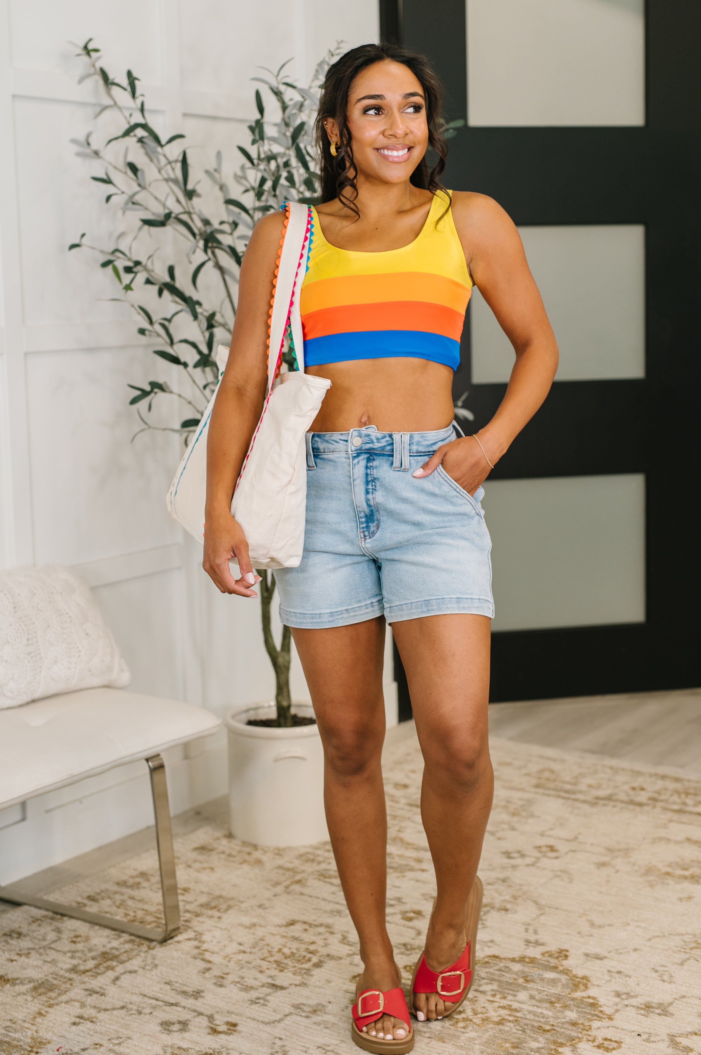 Smiling woman wearing blue, orange, and yellow striped ruched swim crop top with light denim shorts indoors