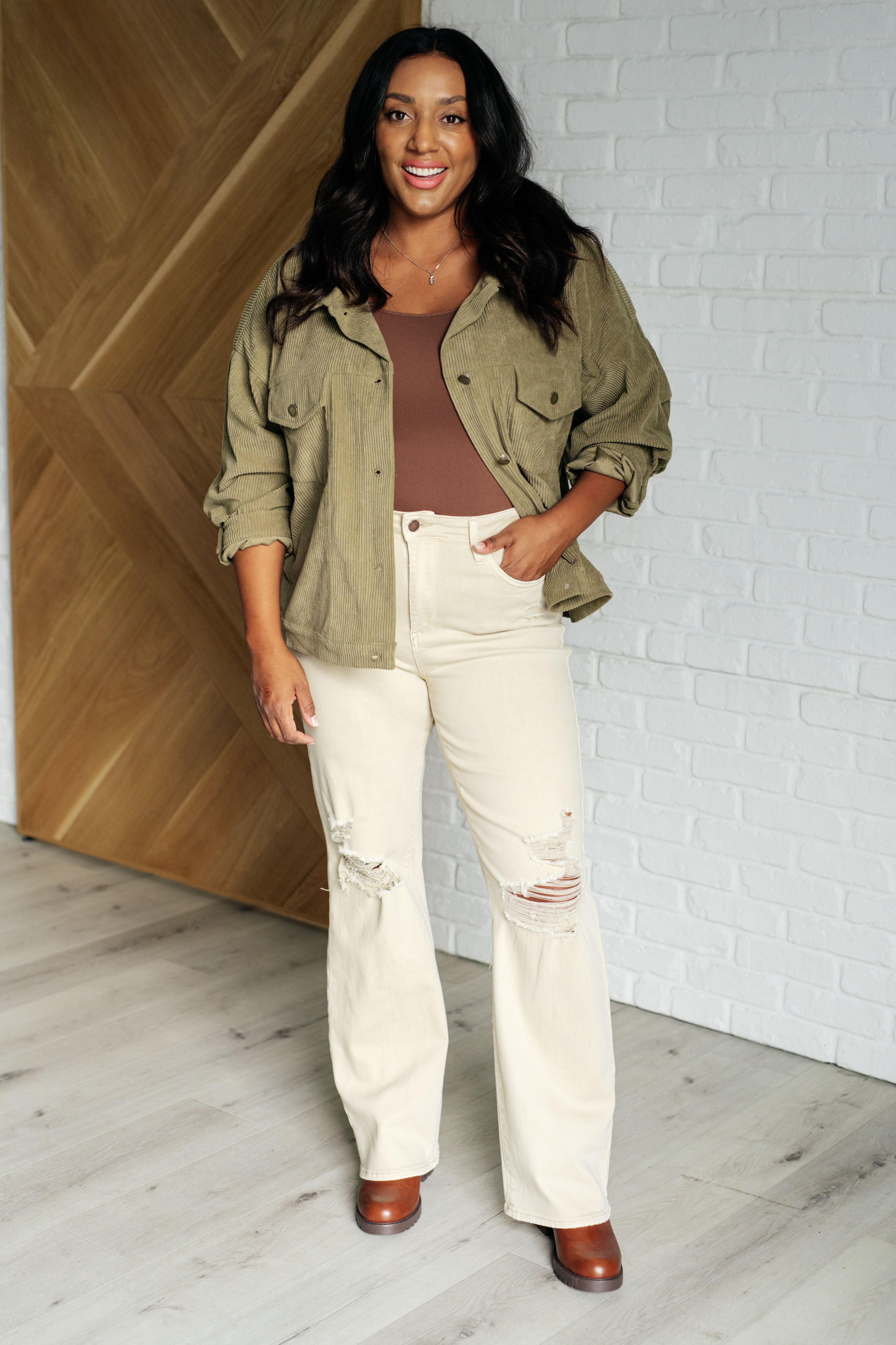 Smiling woman with dark hair wearing an olive corduroy jacket over a brown top and ripped cream jeans indoors