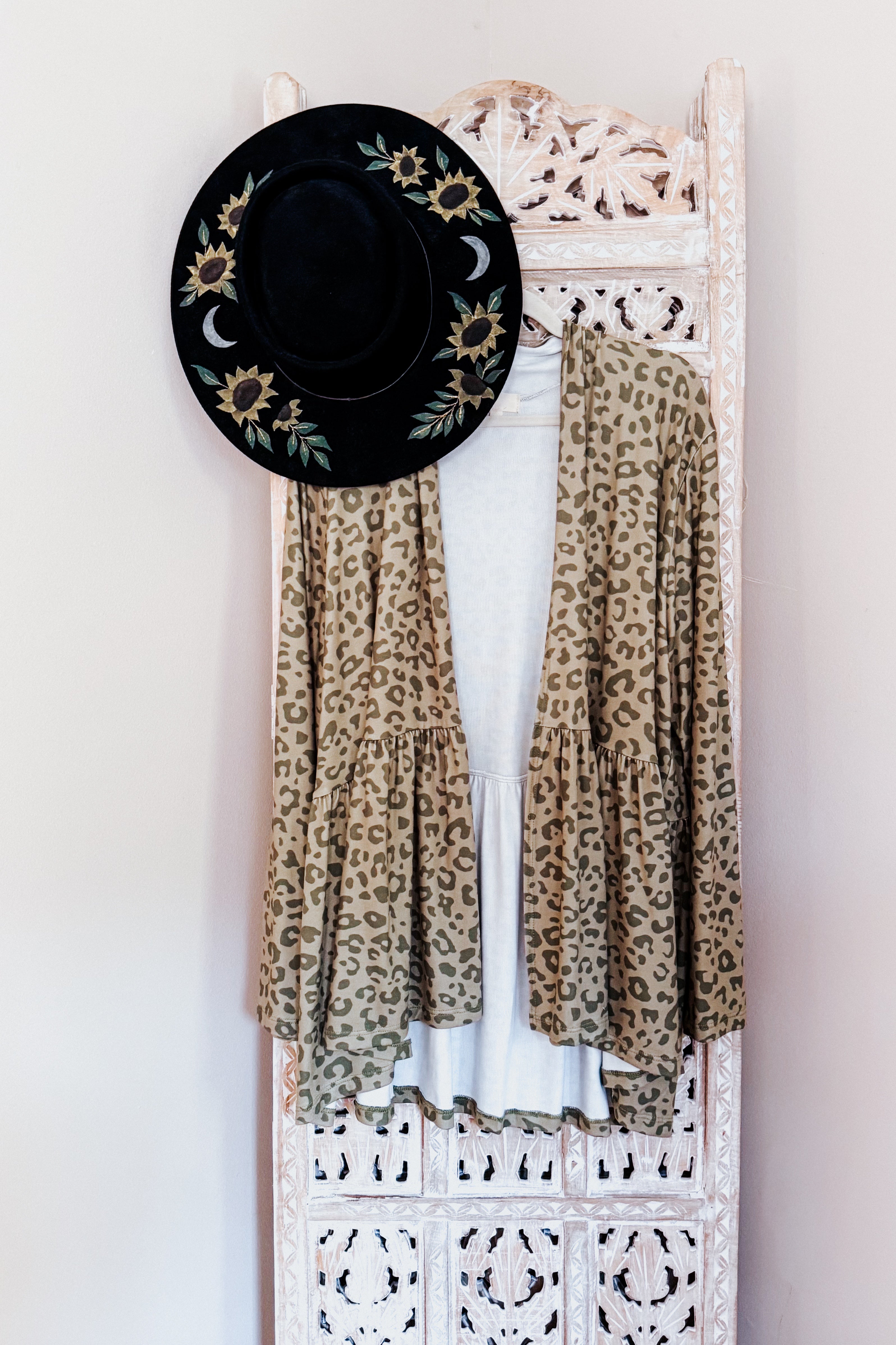 Green and brown leopard print open peplum cardigan hanging with embroidered black hat on carved wood screen in eclectic space