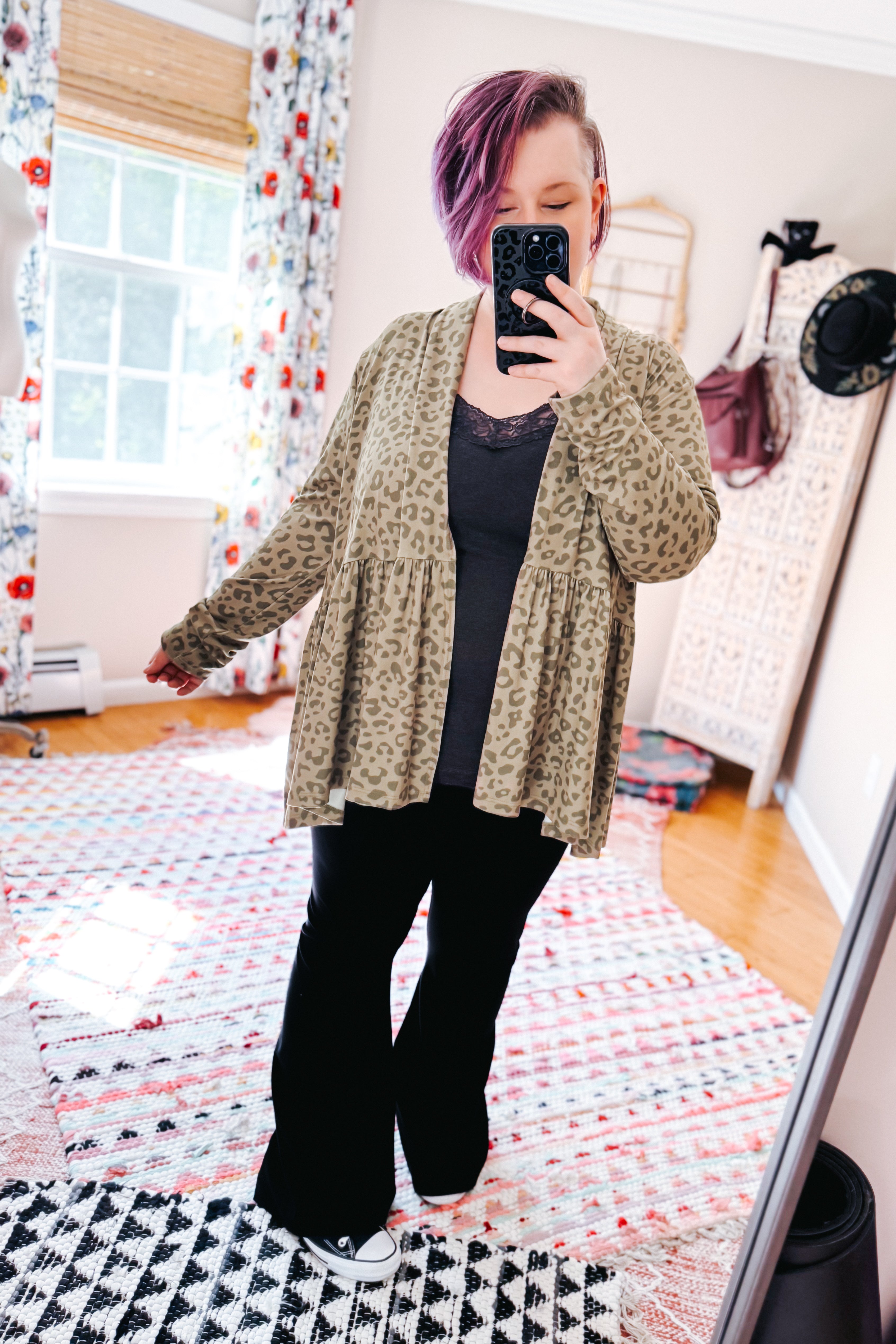 Woman with purple hair wearing green leopard print peplum cardigan over black top and pants in bright eclectic room with patterned rugs