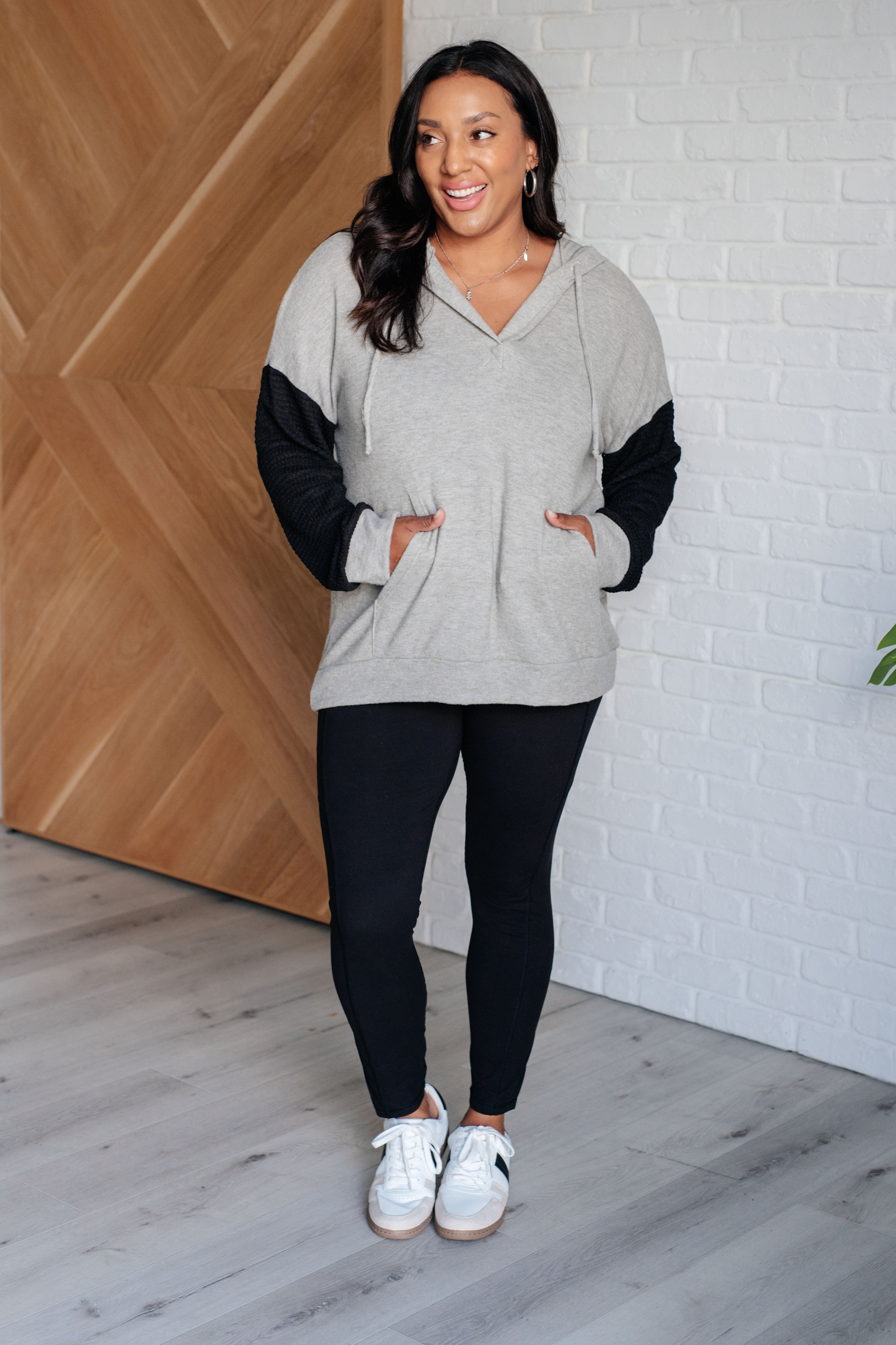 Smiling woman in grey and black color block hoodie paired with black leggings and white sneakers in styled indoor space