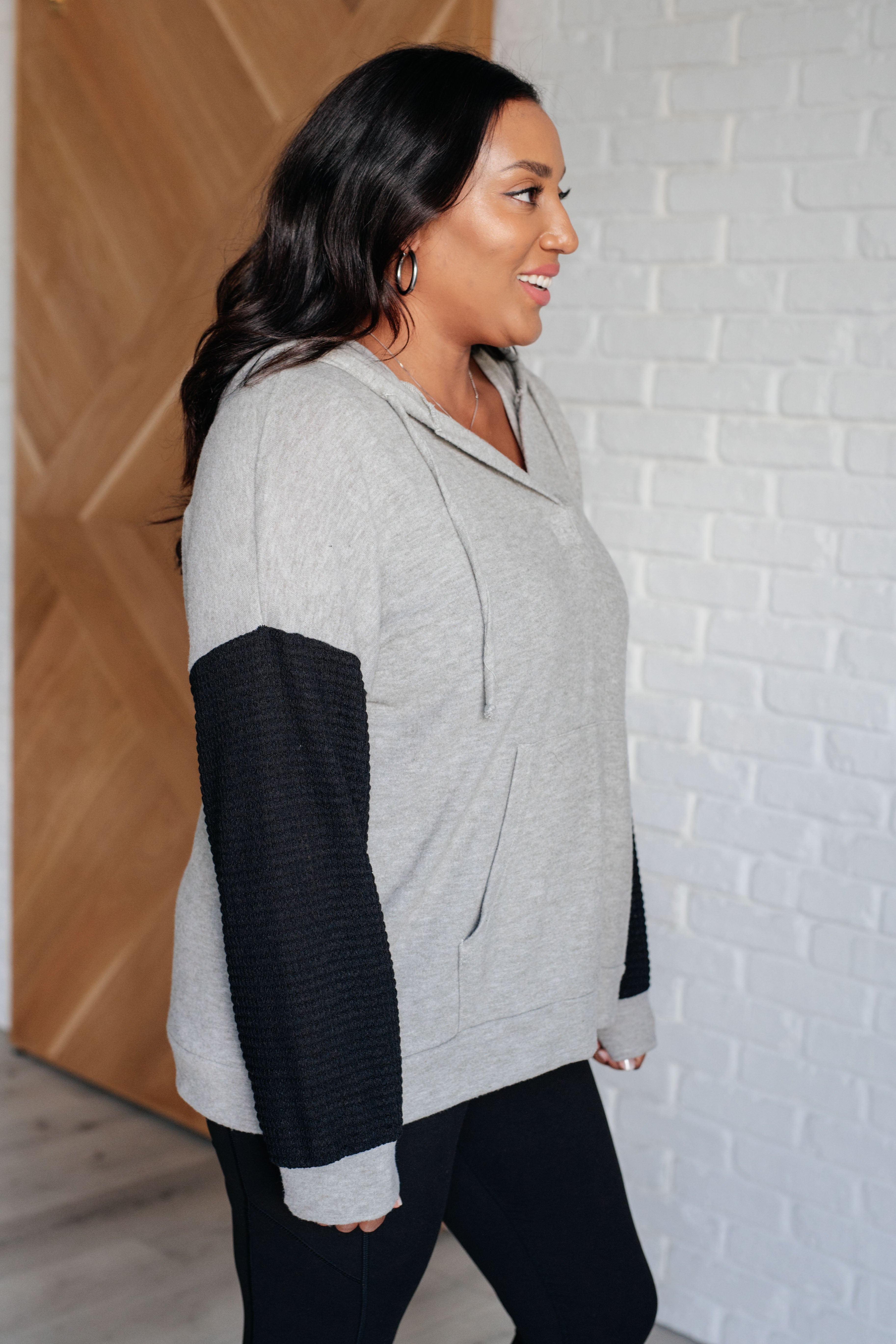 Woman wearing grey hoodie with contrasting textured black sleeves and black pants standing indoors by wood and white walls
