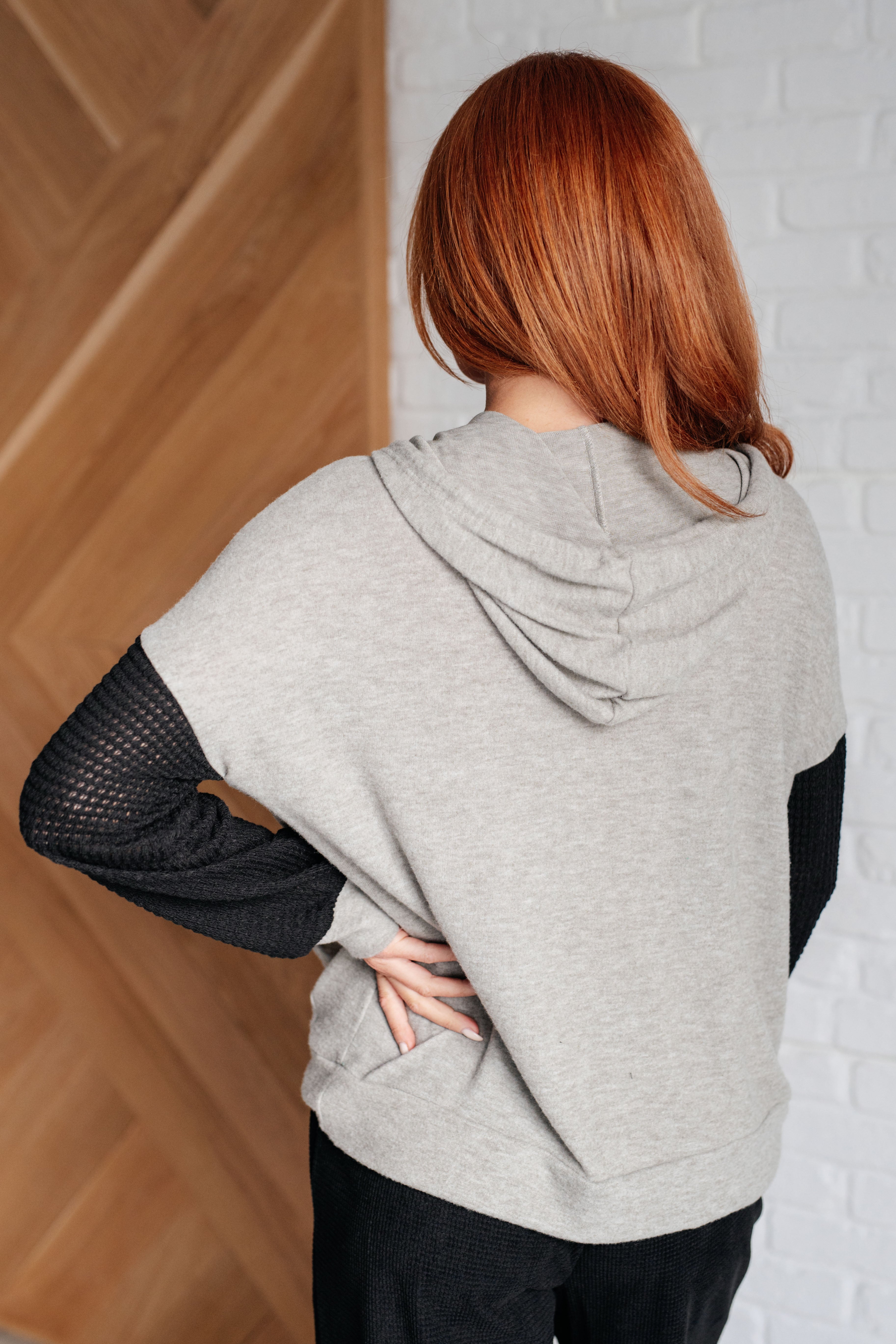 Woman with red hair wearing grey hoodie with black textured color block sleeves, standing indoors with hand on hip