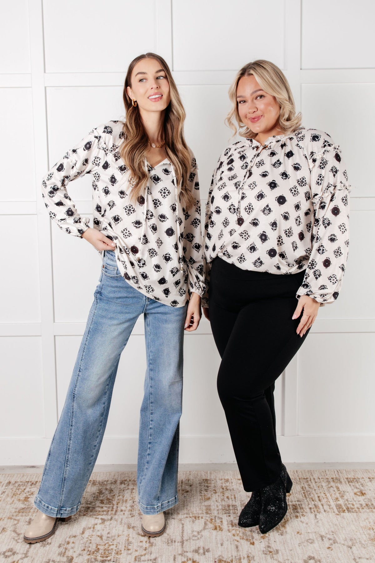 Two women model white floral blouses with ruffle details styled with blue jeans and black pants in a bright interior