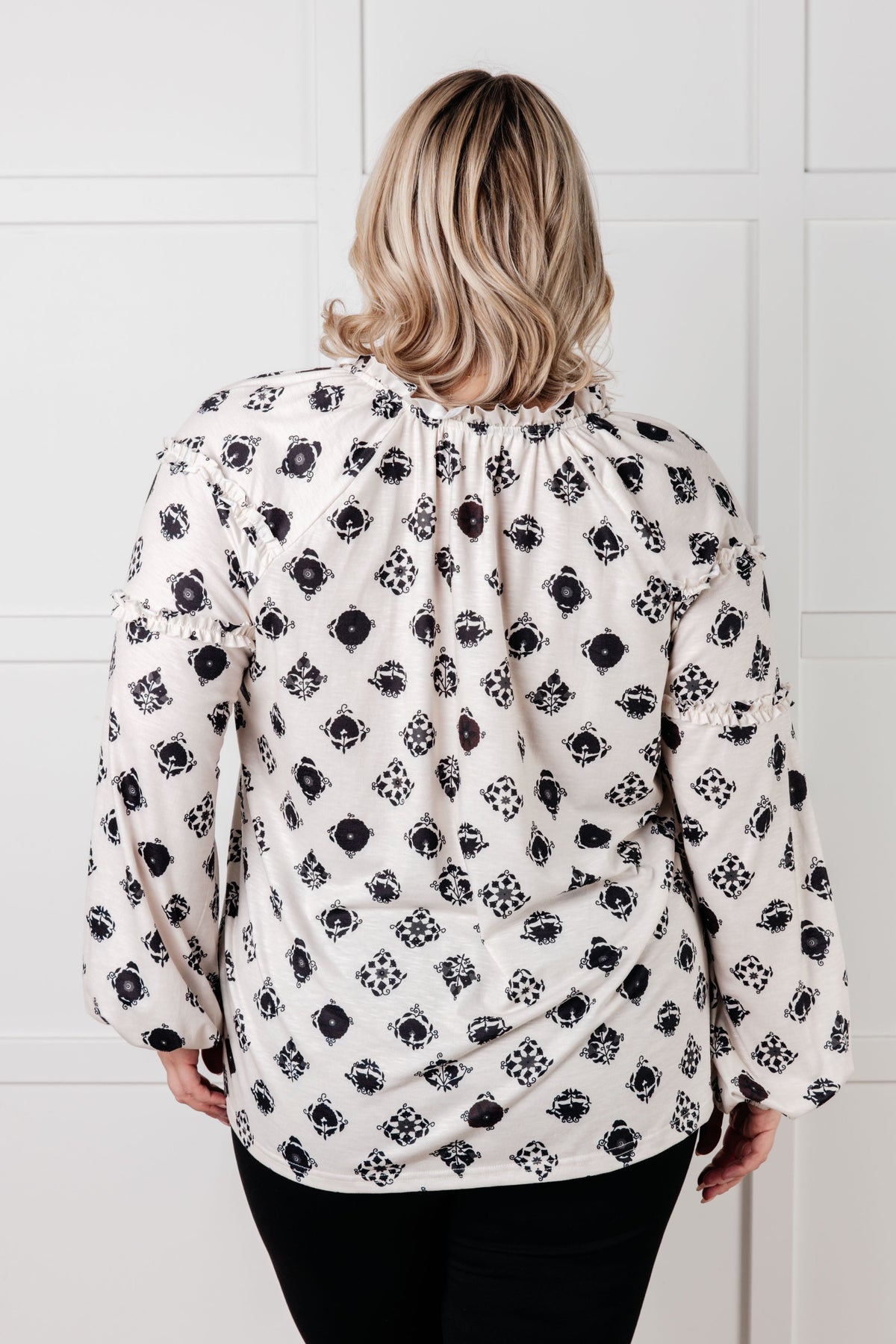 Back view of a woman wearing a white floral tile blouse with a split neckline and balloon sleeves styled with black pants in a bright interior