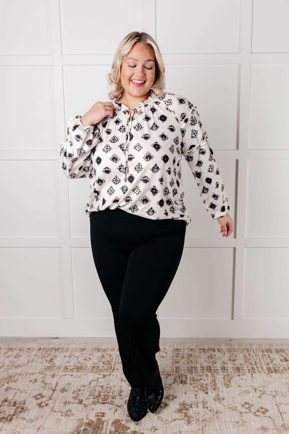 Smiling woman wearing a white floral tile blouse with ruffle details and black pants in a bright paneled room