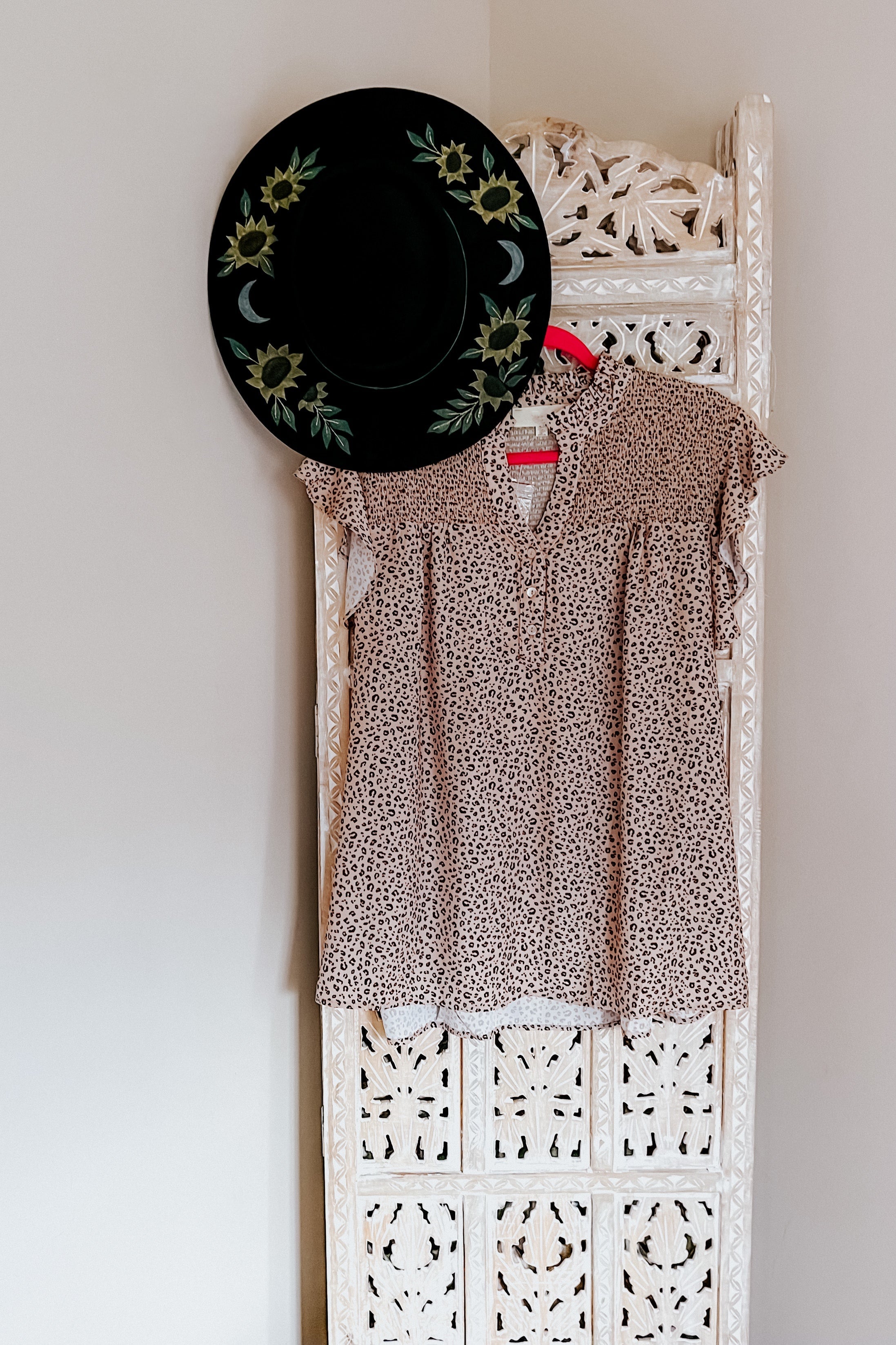 Taupe animal print blouse with flutter sleeves hanging on ornate white carved wood panel with black hat decorated with sunflowers