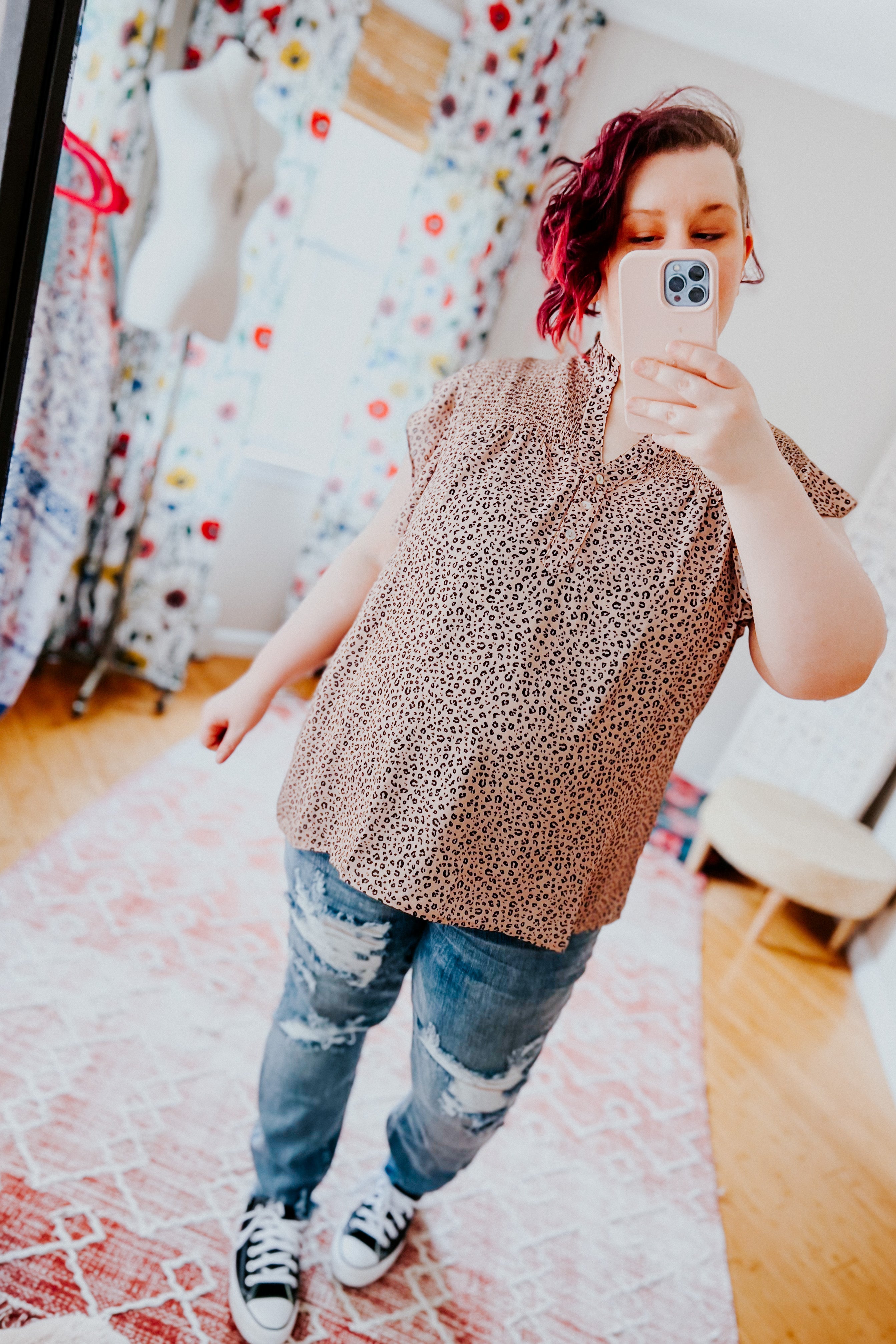 Woman wearing taupe animal print sleeveless top paired with distressed jeans and black sneakers in bright room with floral curtains