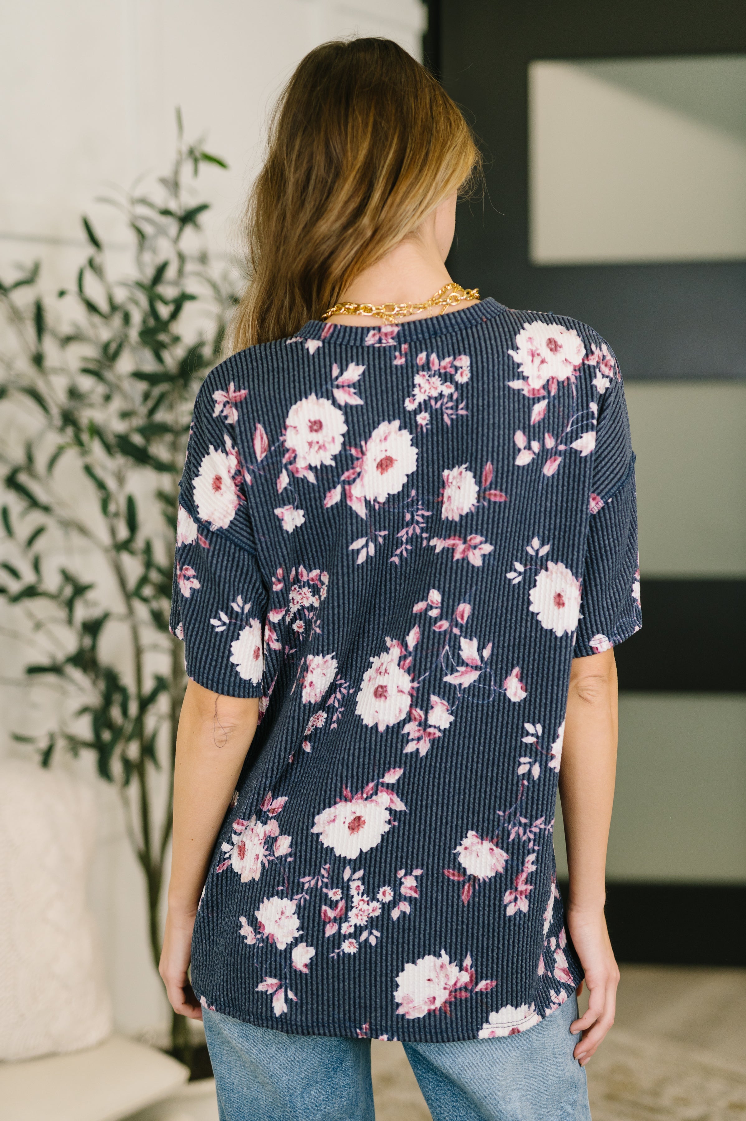 Back view of woman wearing navy floral textured short sleeve top paired with light blue jeans indoors