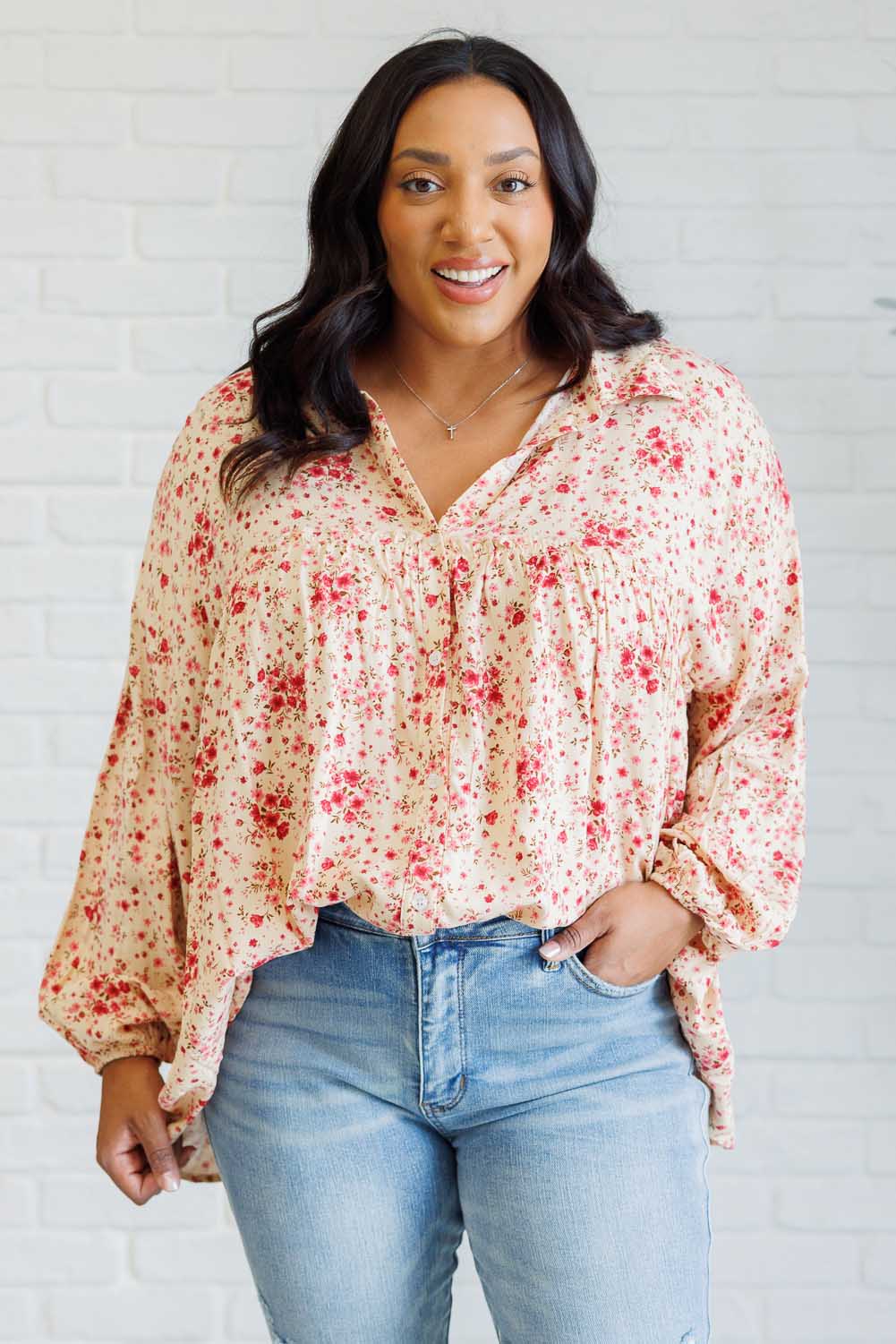 Smiling woman wearing a pink floral button down shirt with balloon sleeves and jeans indoors against a white brick wall