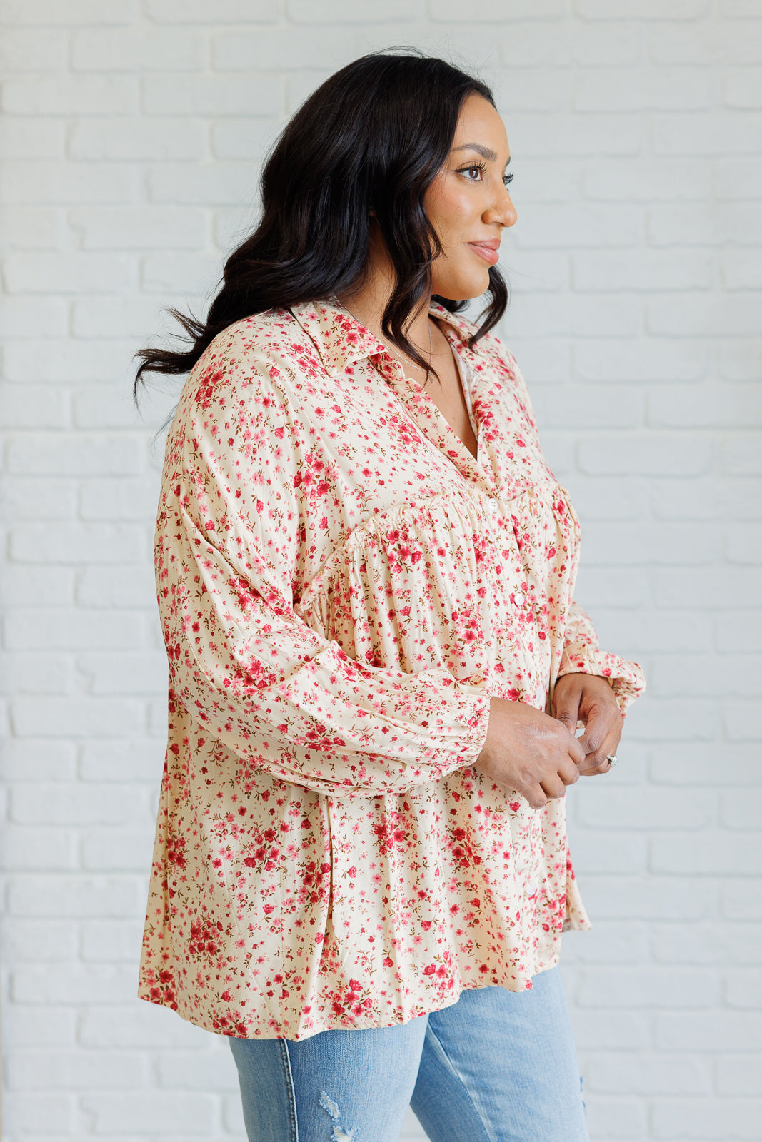 Plus size woman modeling a pink floral button down shirt with balloon sleeves and light wash jeans against a white brick wall indoors