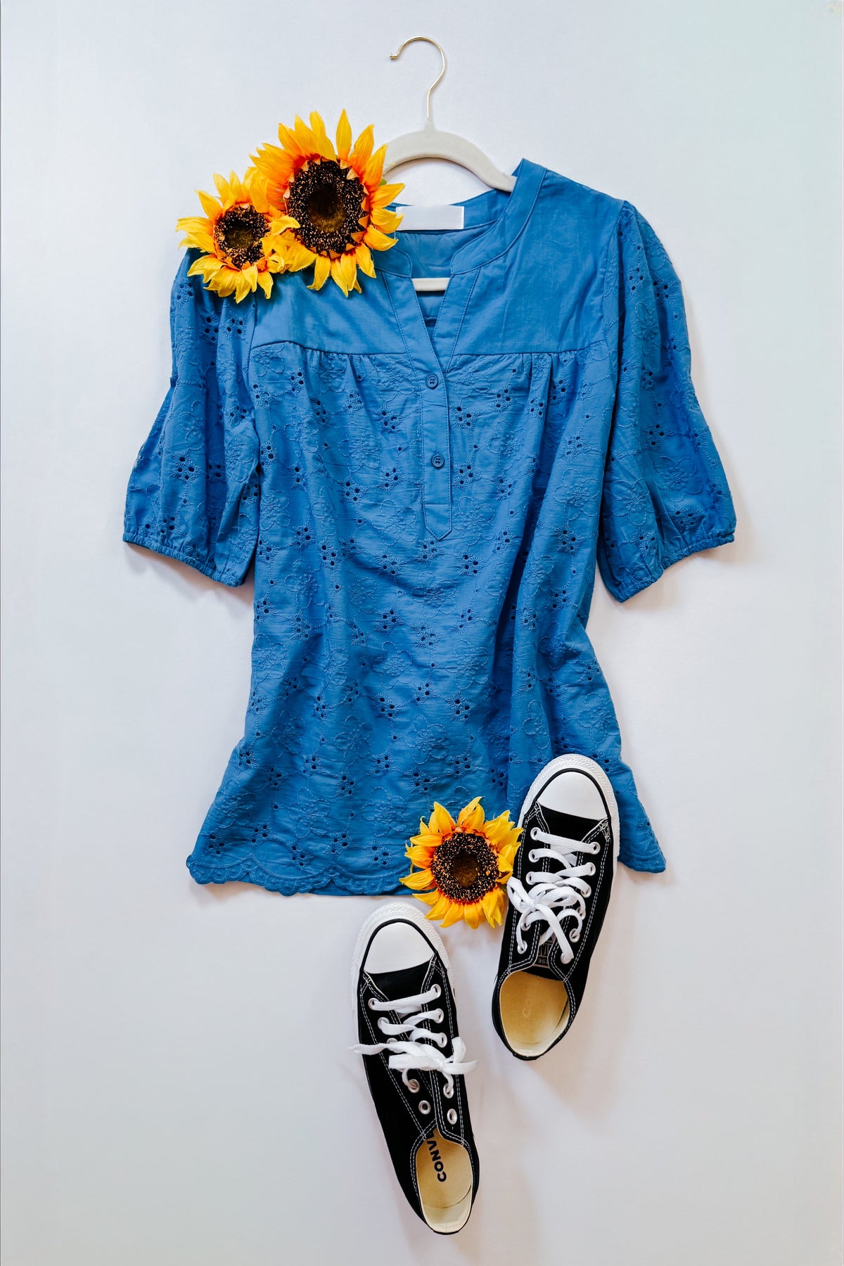 Blue eyelet blouse with three quarter sleeves styled with sunflowers and black lace up sneakers on a white background