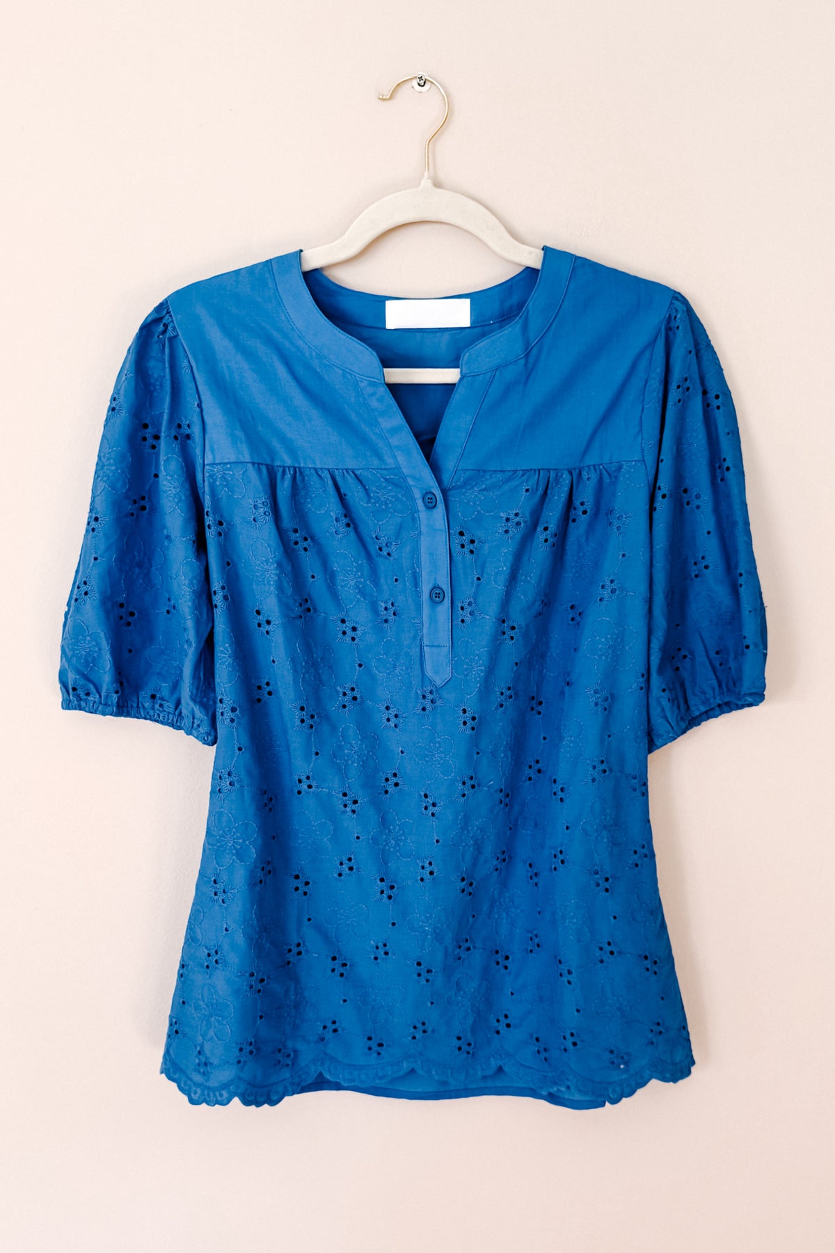 Blue eyelet blouse hanging on a white hanger with a split neckline button placket puff sleeves and scalloped hem