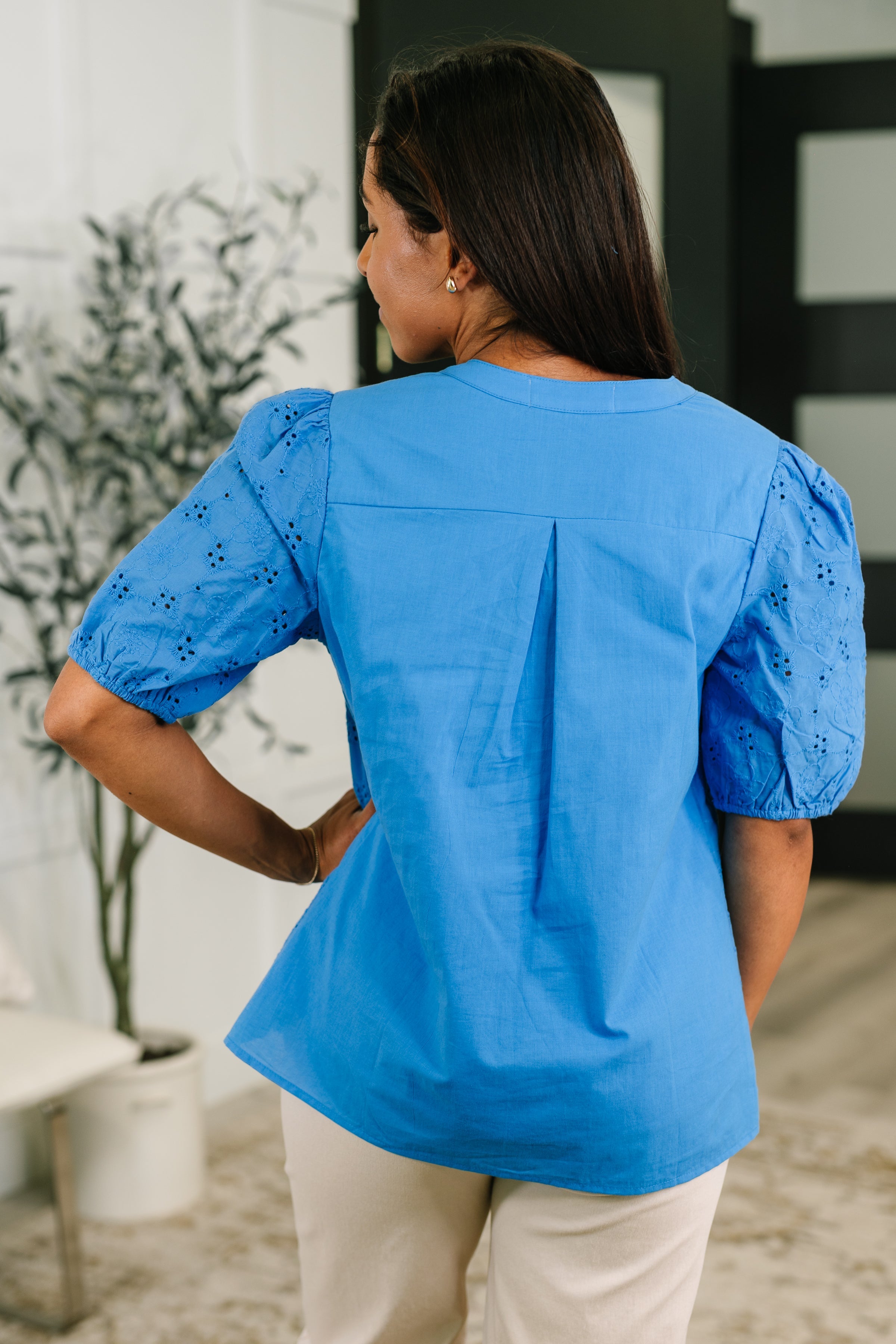Woman wearing a blue eyelet blouse with puff sleeves and back pleat in a bright eclectic and size inclusive indoor space