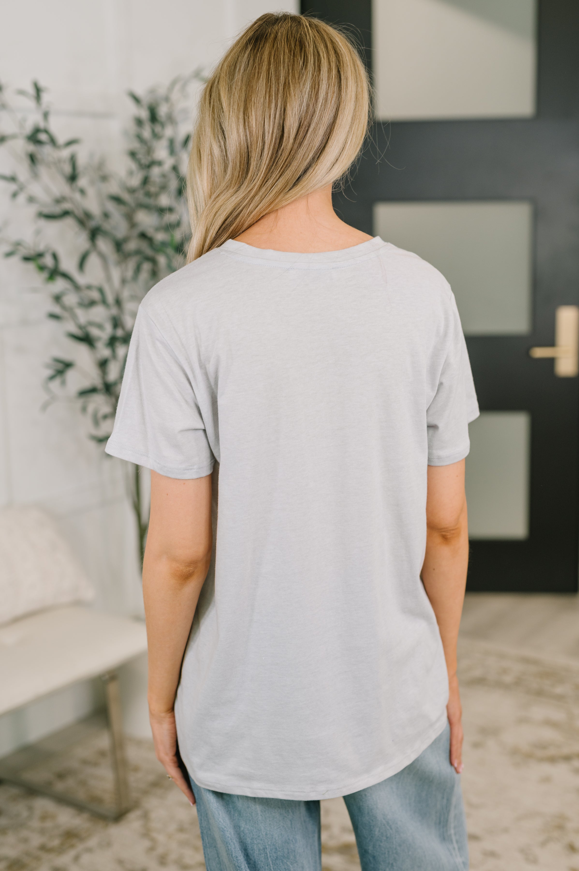 Back view of woman wearing heather grey relaxed fit tee with short sleeves and light-wash jeans in cozy home setting