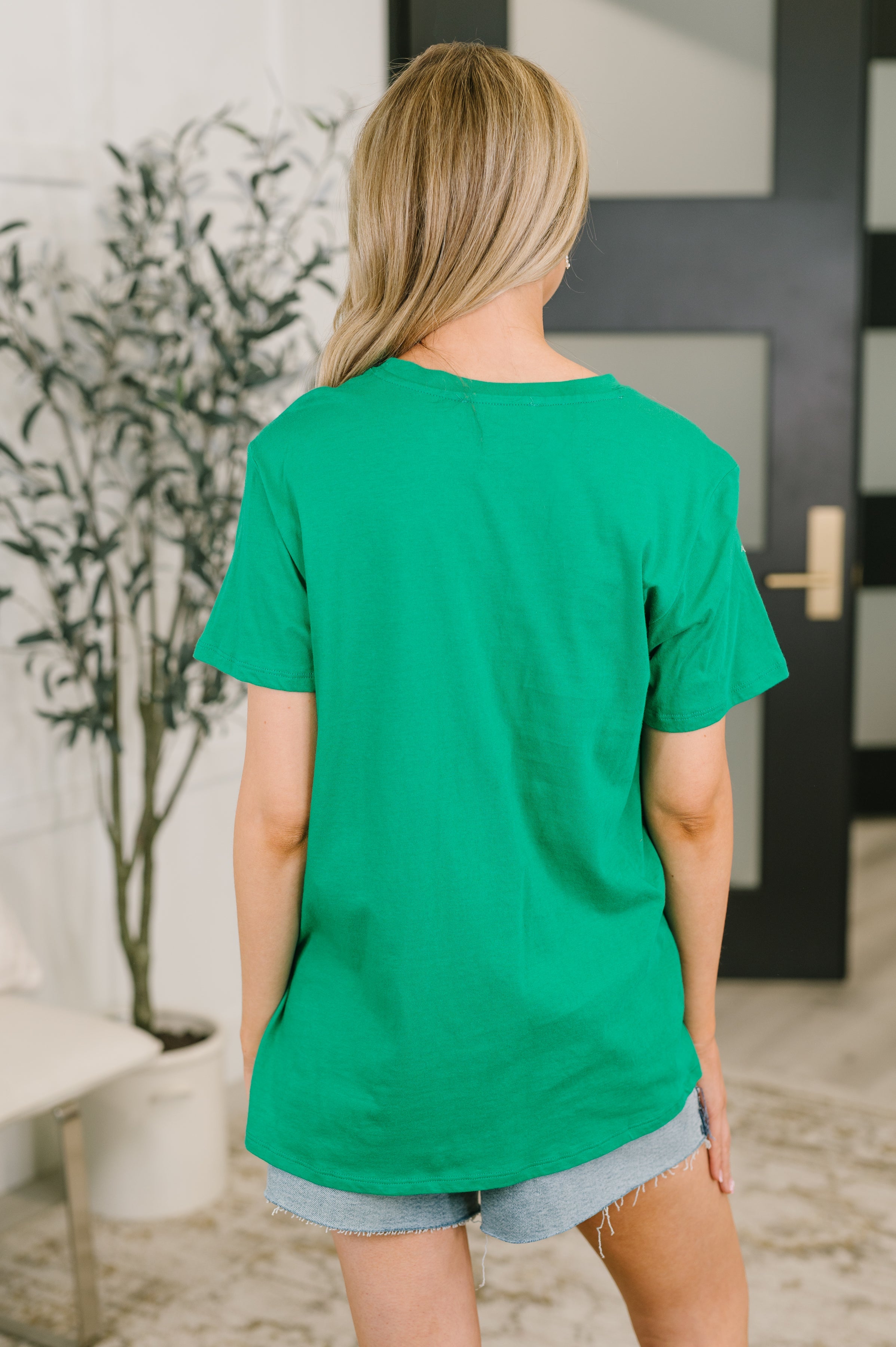 Back view of person wearing Kelly Green relaxed fit tee paired with casual light denim shorts in bright indoor room