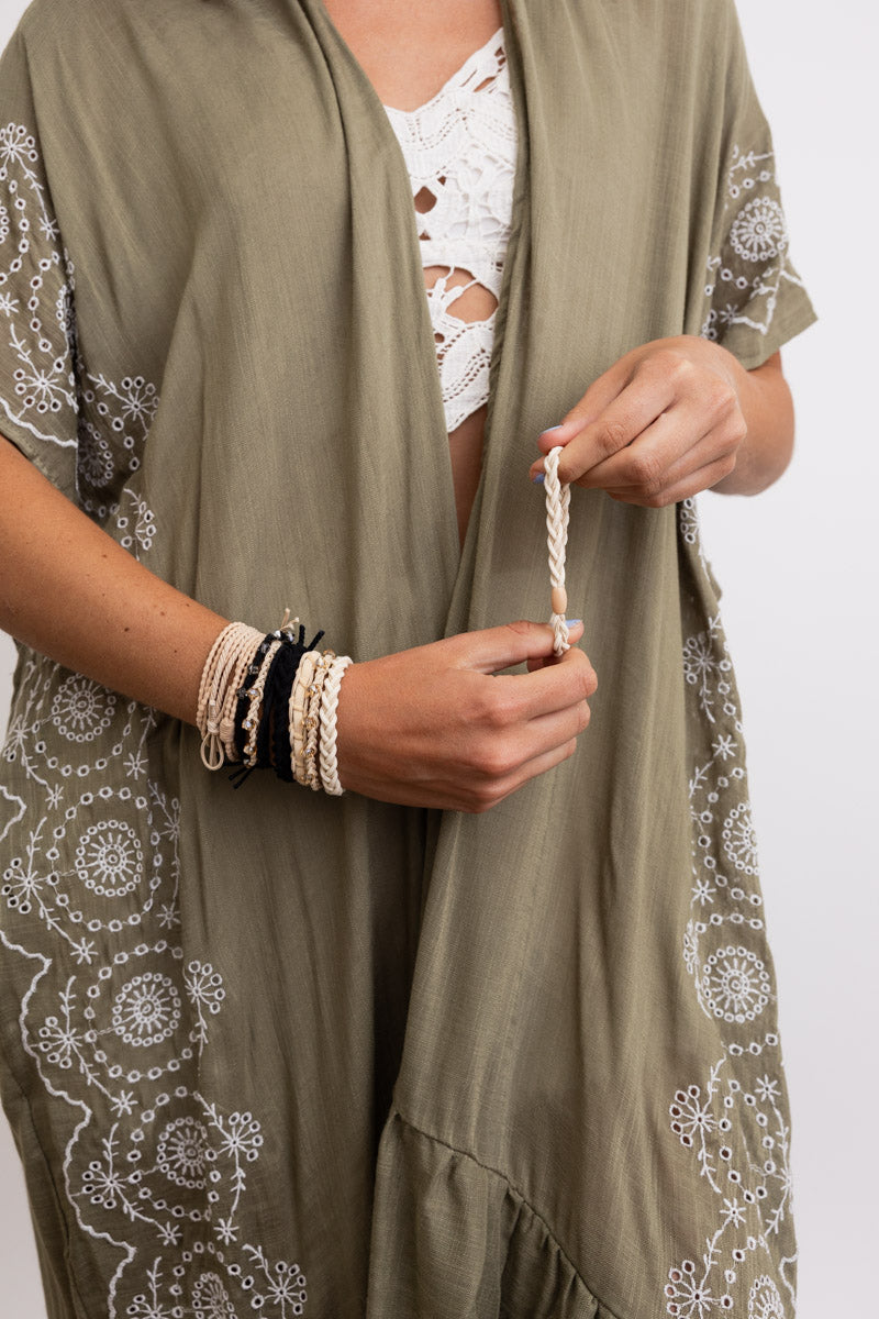 Close-up of person wearing cream and black stackable hair tie bracelets paired with olive green embroidered cover-up and white lace top